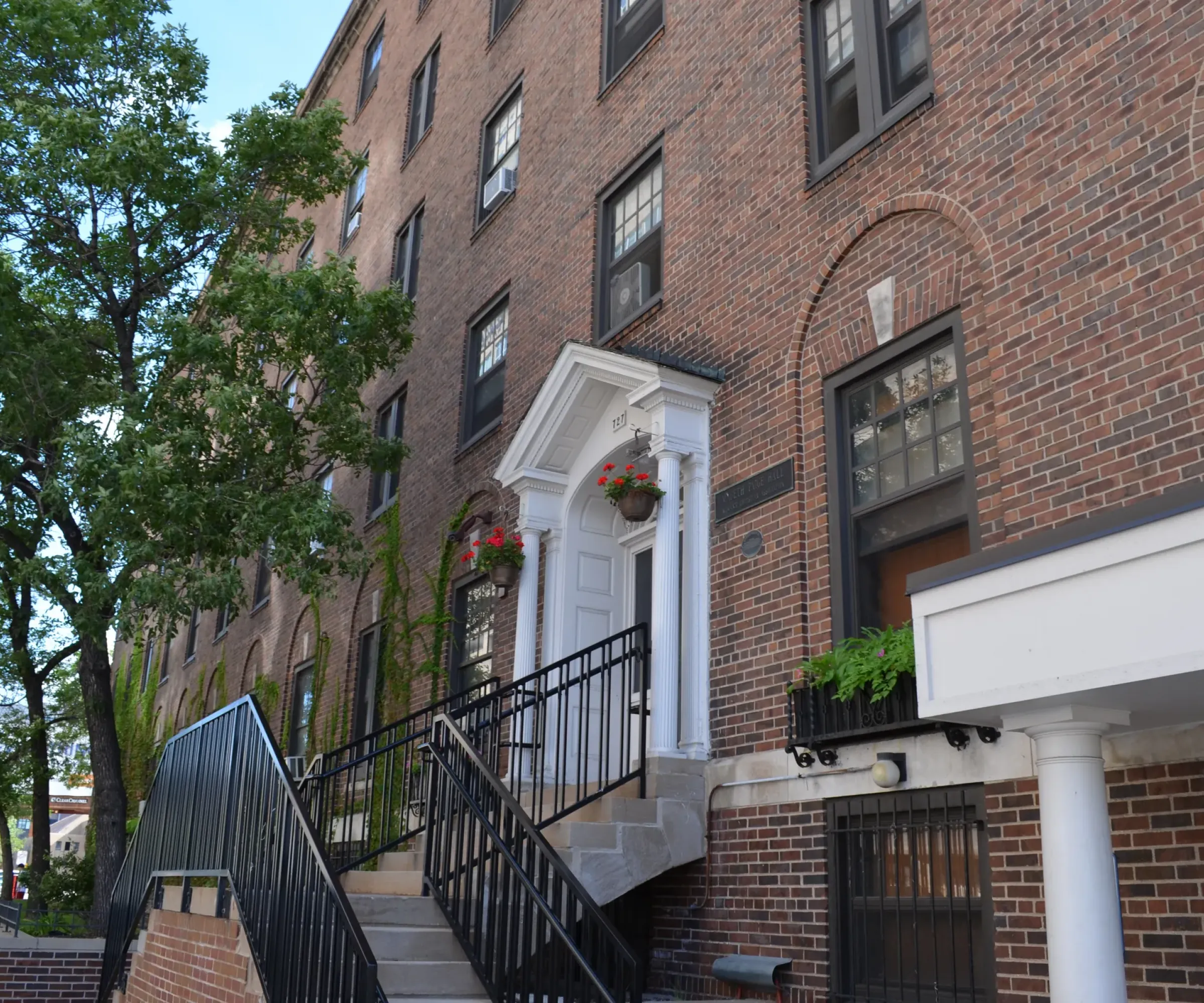 Aeon Affordable Homes and Community Affordable apartment homes with brick exteriors, stairs, black railings, and red flowers in hanging pots near a large tree.