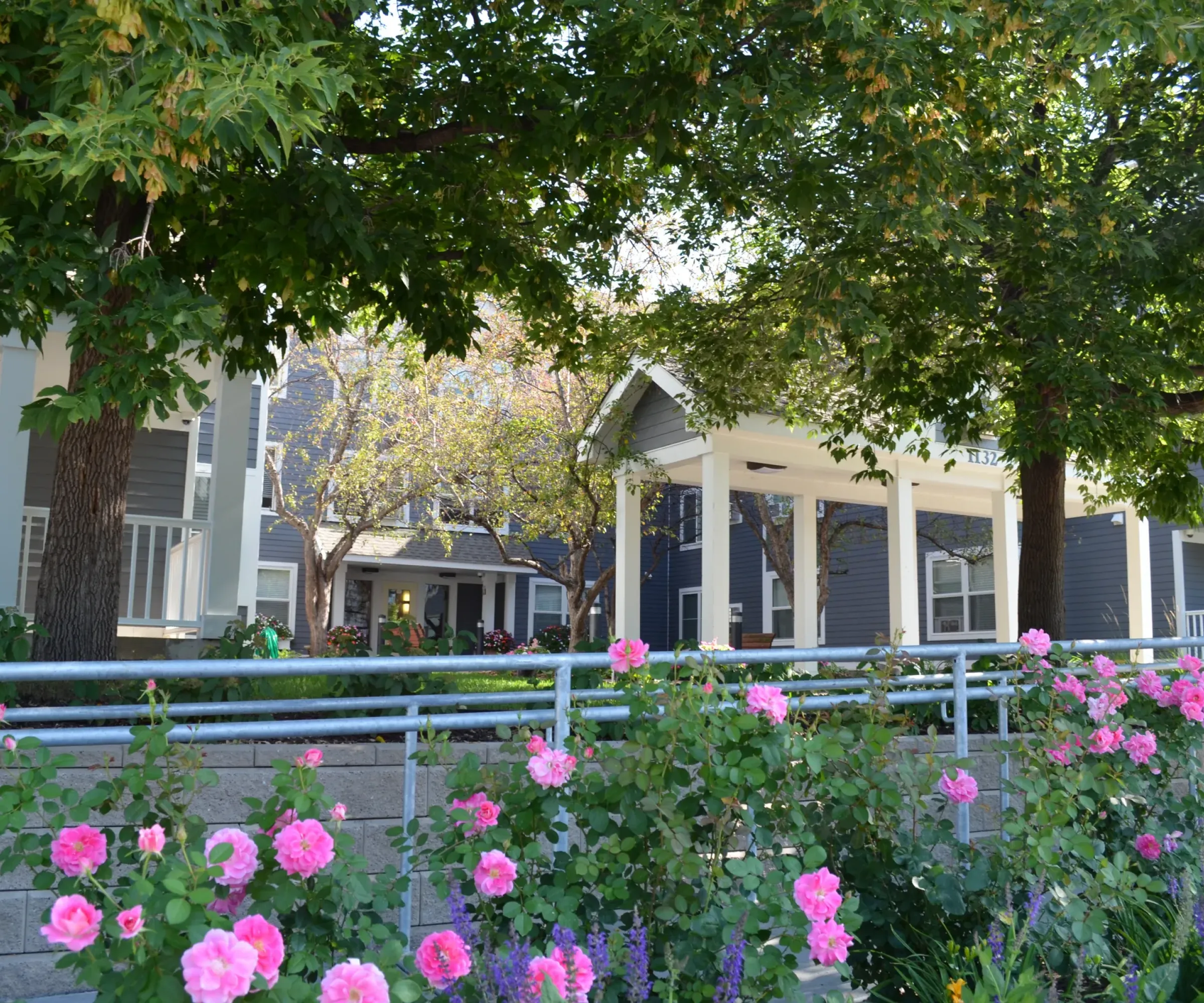 Aeon Affordable Homes and Community A charming affordable apartment home with a porch and lush garden, featuring pink roses and leafy trees in the foreground.