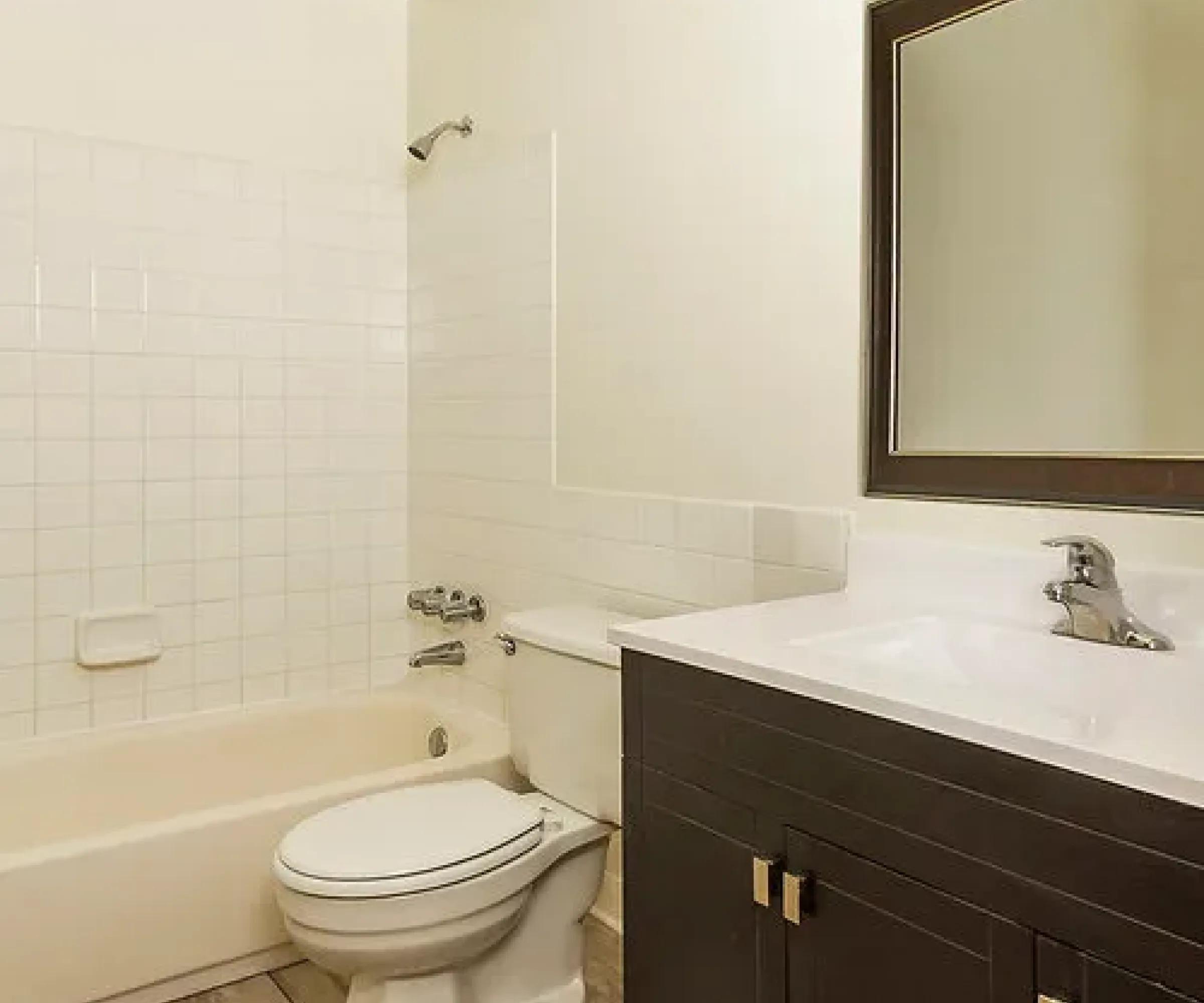 Aeon Affordable Homes Cobblestone Court Apartments feature a bathroom with a bathtub, toilet, dark wood vanity, sink, large mirror, and tiled floor and walls.