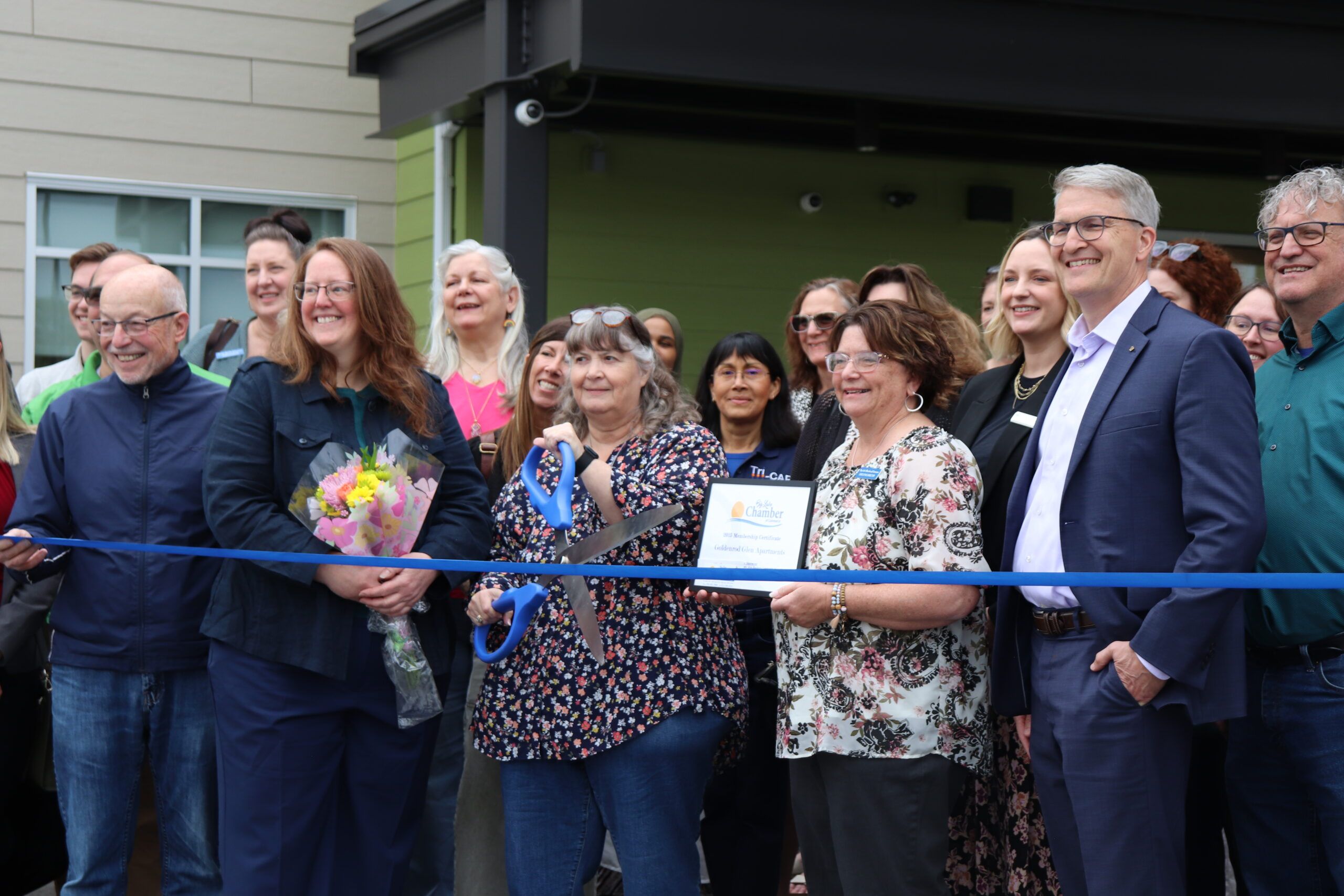 Aeon Affordable Homes A group of people at a ribbon-cutting ceremony with a woman holding large scissors and others smiling.