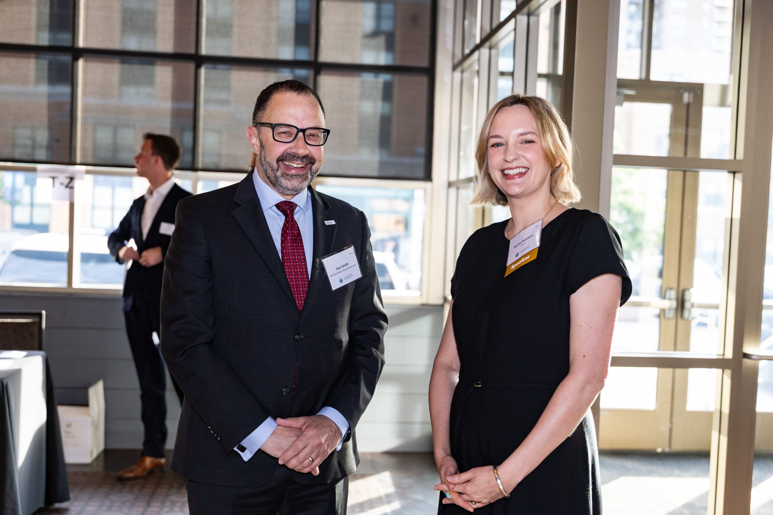 Aeon Affordable Homes Two people in business attire smile at the camera at an indoor event with sunlight streaming through large windows.