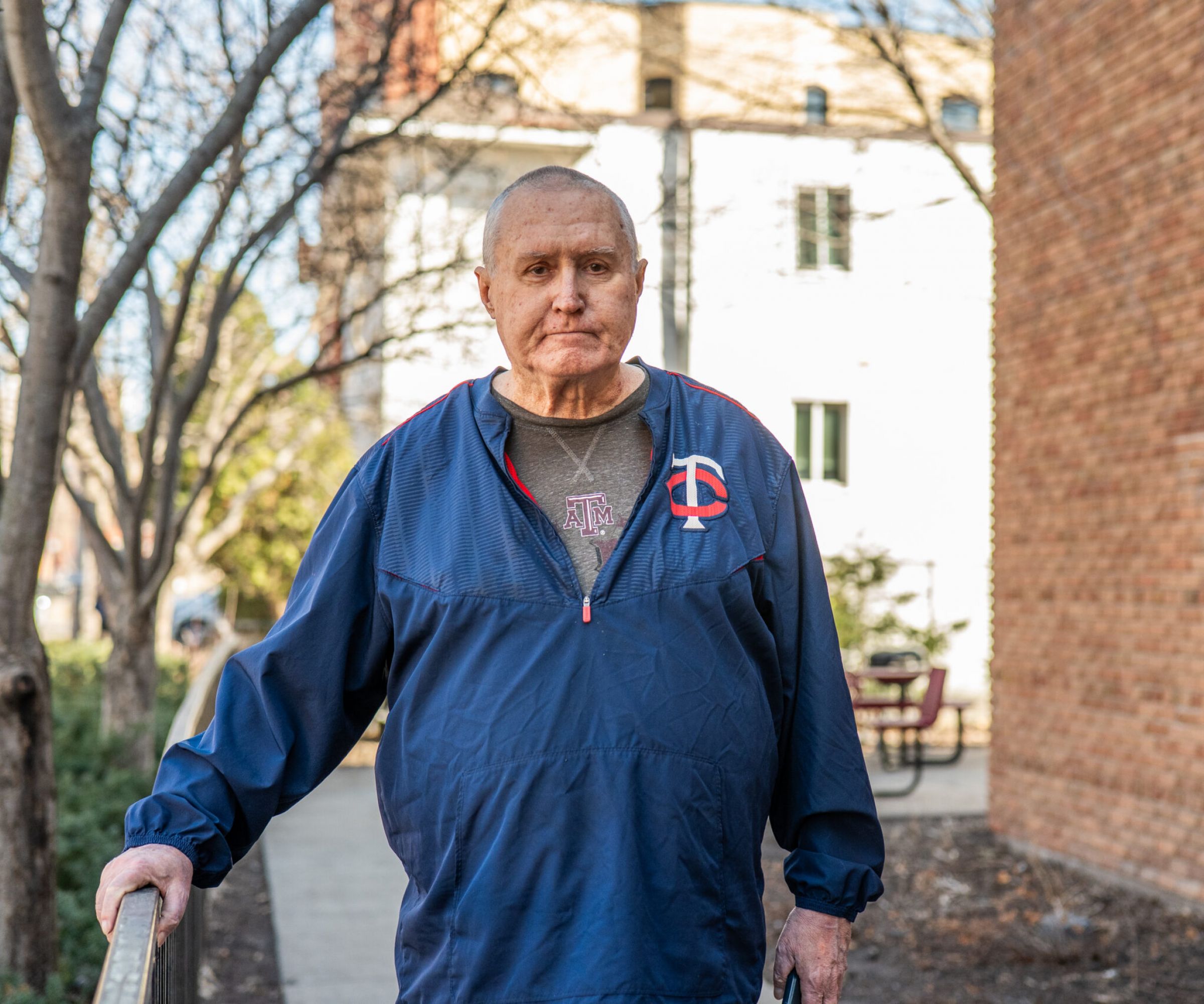 Aeon Affordable Homes An older man in a blue jacket stands outside on a sidewalk—perhaps it's part of Rick's Story—near brick buildings and leafless trees.