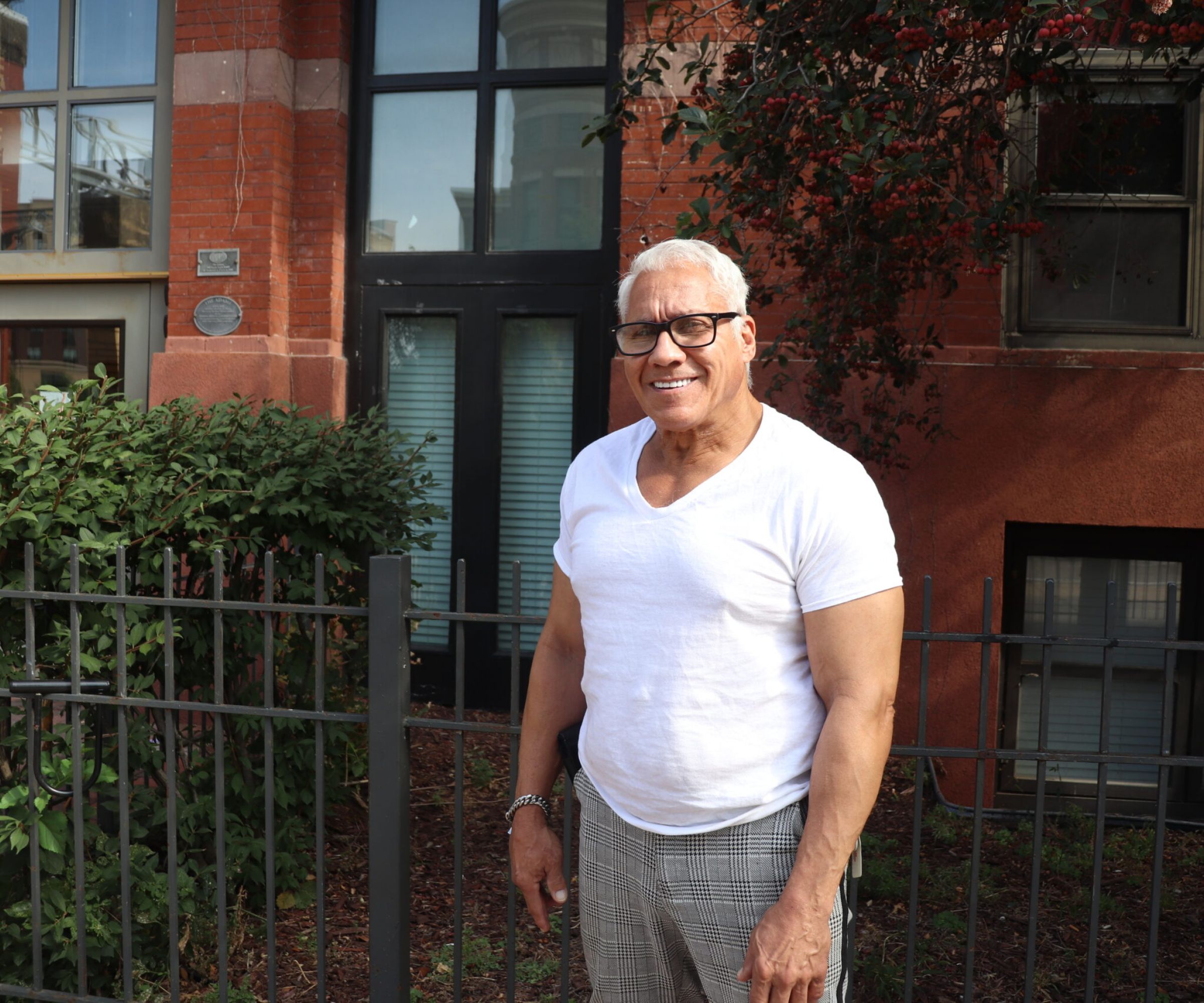 Aeon Affordable Homes Smiling man in glasses and white shirt stands by a fence in front of a brick building with large windows.