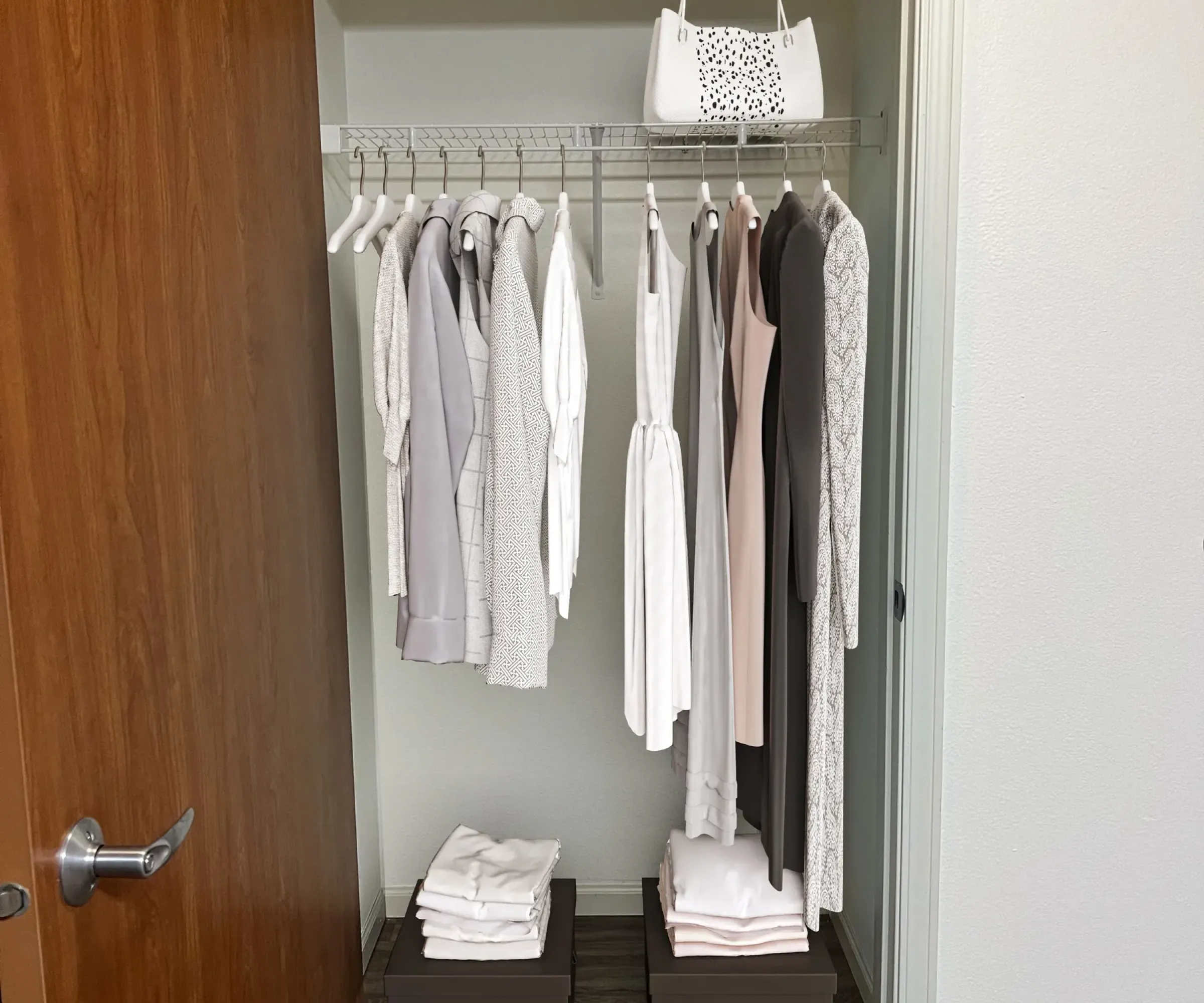 Aeon Affordable Homes and Community Minimalist closet in affordable apartment homes with neutral-toned clothes on hangers, folded items on shelves, and a white handbag atop.