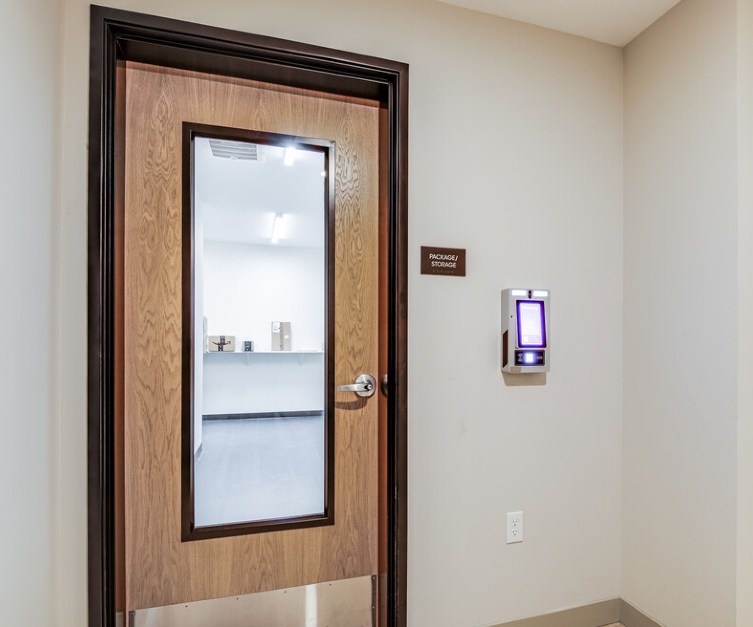 Aeon Affordable Homes and Community Wooden door with a glass panel leads to "Packaging Storage." A control panel is mounted beside it, typical of affordable apartment homes.