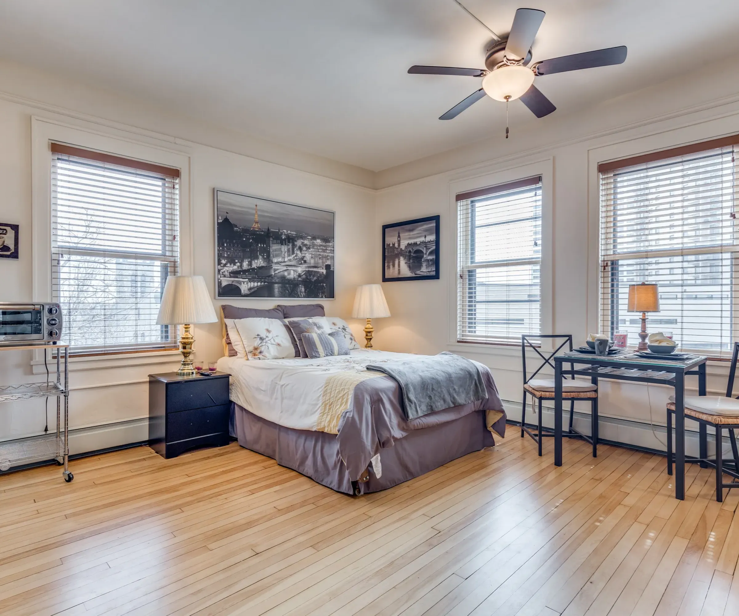 Aeon Affordable Homes and Community A cozy bedroom in affordable apartment homes, with a bed, small dining table, artwork, and ceiling fan on wooden floors beneath large windows.