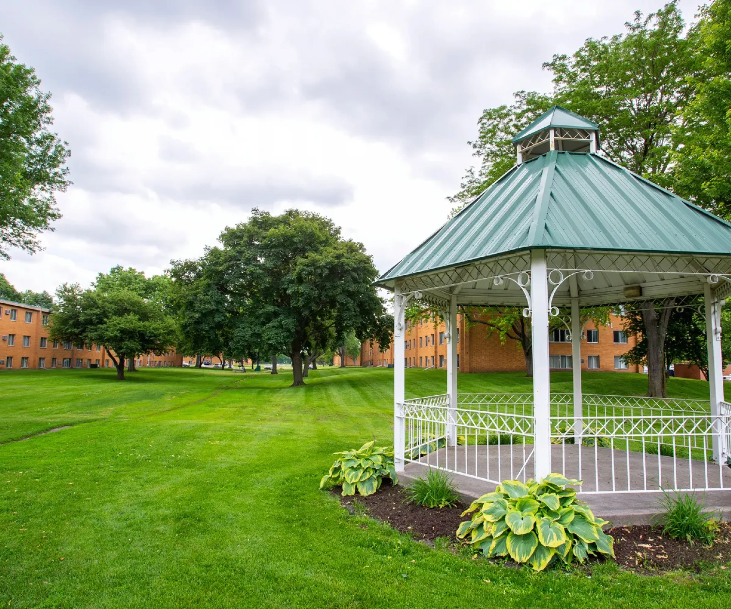 Aeon Affordable Homes A white gazebo on a lush green lawn with trees and brick buildings in the background under a cloudy sky.