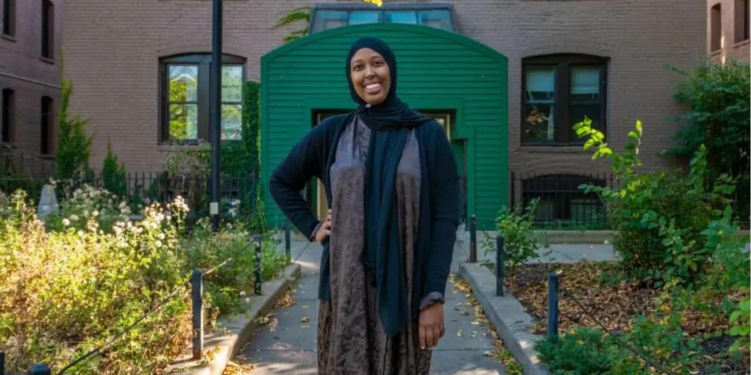 Aeon Affordable Homes and Community with a smiling woman standing outside a brick apartment building surrounded by green plants and trees.