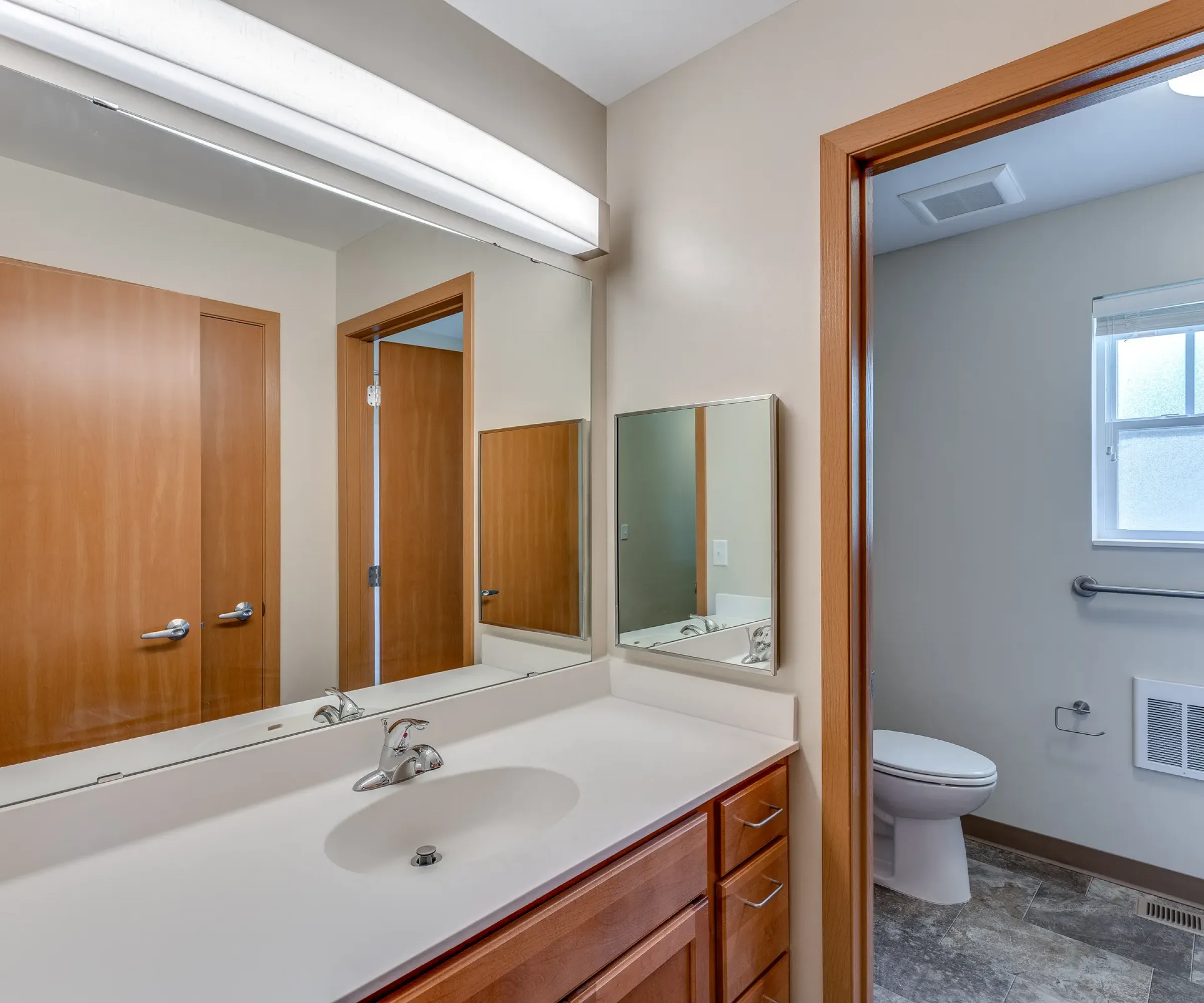 Aeon Affordable Homes and Community Modern bathroom in affordable apartment homes, featuring a wooden vanity, sink, mirror, toilet, open door, and window.