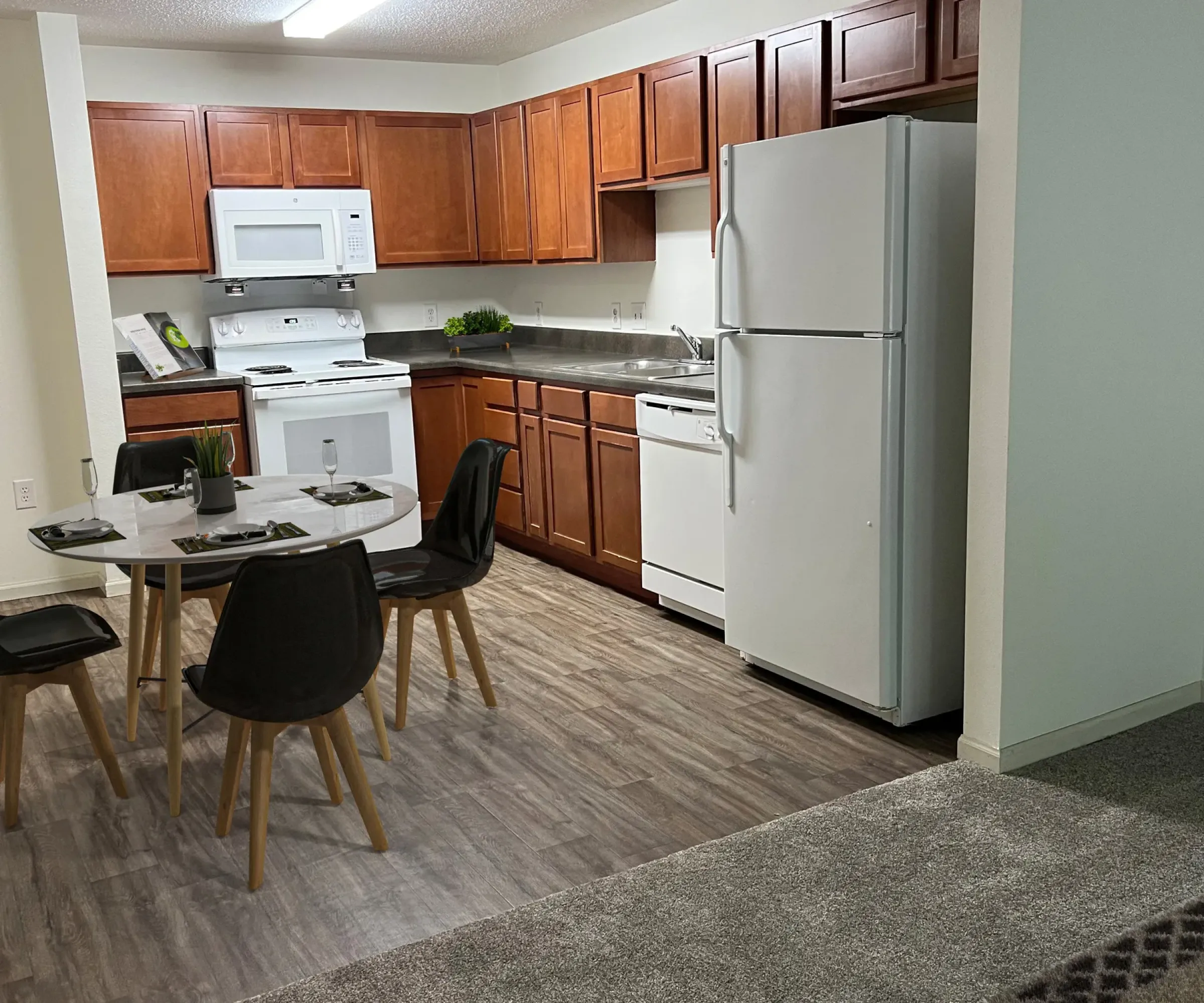 Aeon Affordable Homes and Community An affordable apartment home kitchen with wooden cabinets, white appliances, and a dining table for four on a wood-patterned floor.