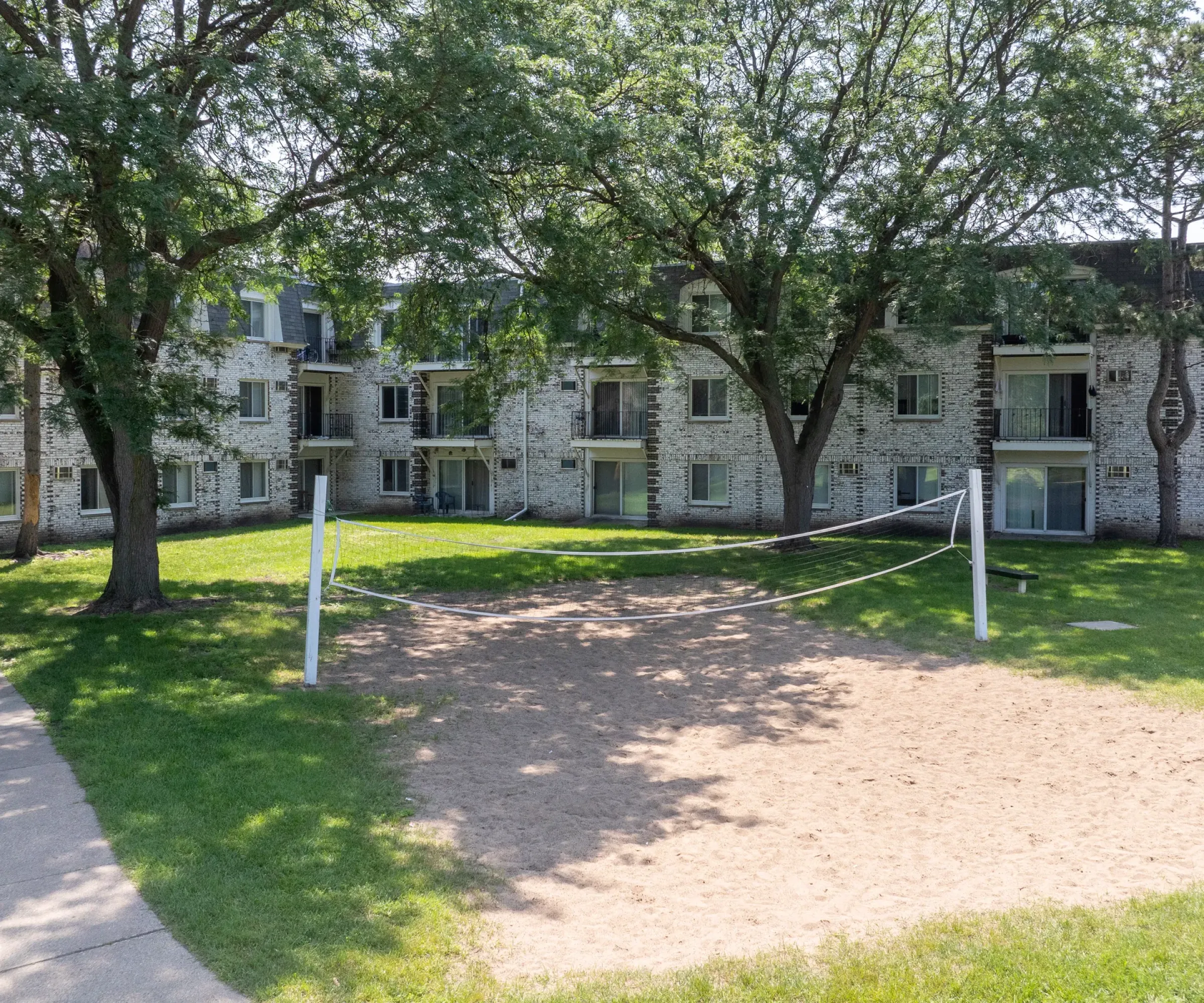 Aeon Affordable Homes A sandy volleyball court with a net graces Blooming Meadows, set in front of a brick apartment complex surrounded by trees.
