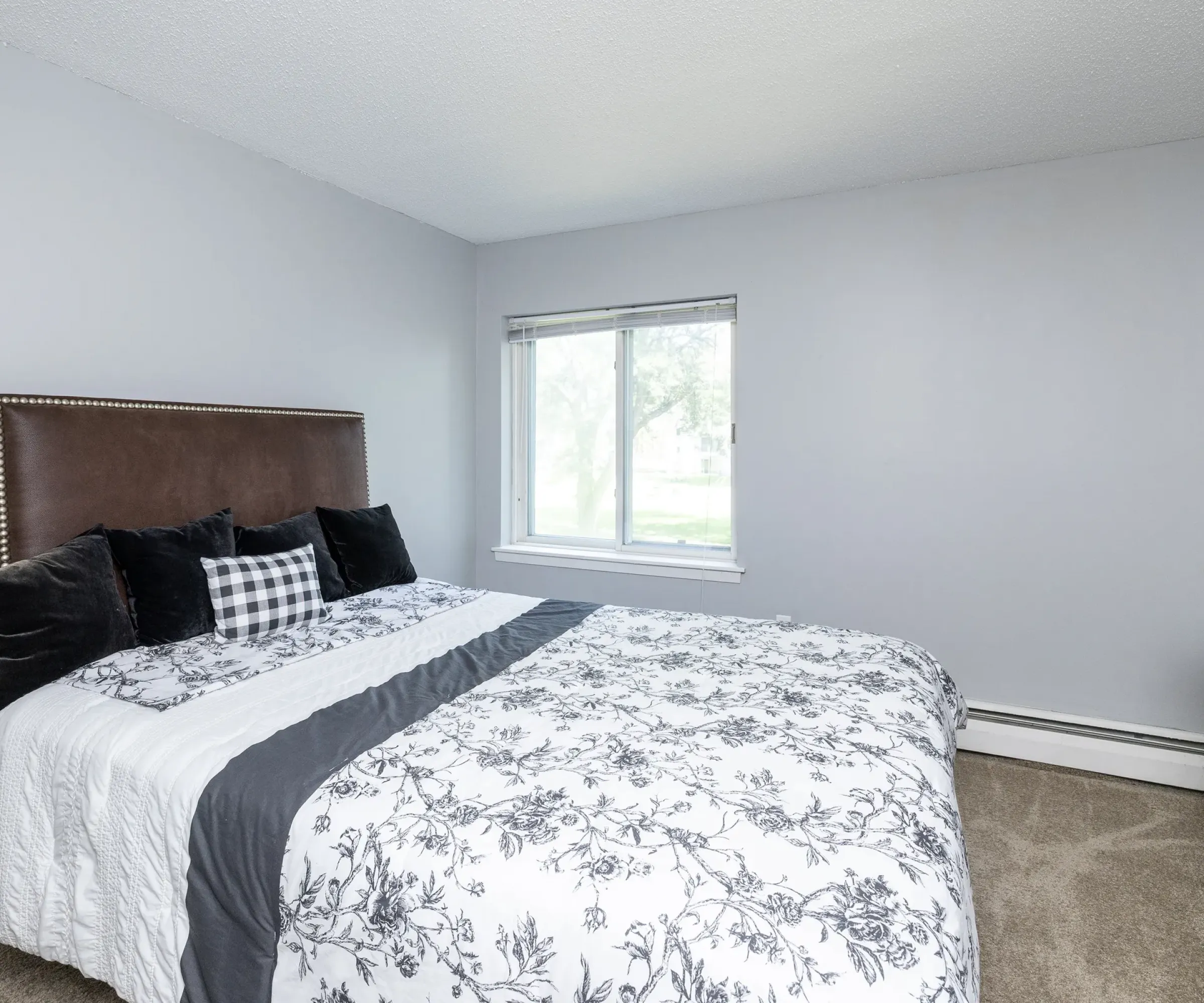 Aeon Affordable Homes A cozy Blooming Meadows bedroom with a floral-patterned bedspread, dark pillows, a window, and a dresser stacked with books.
