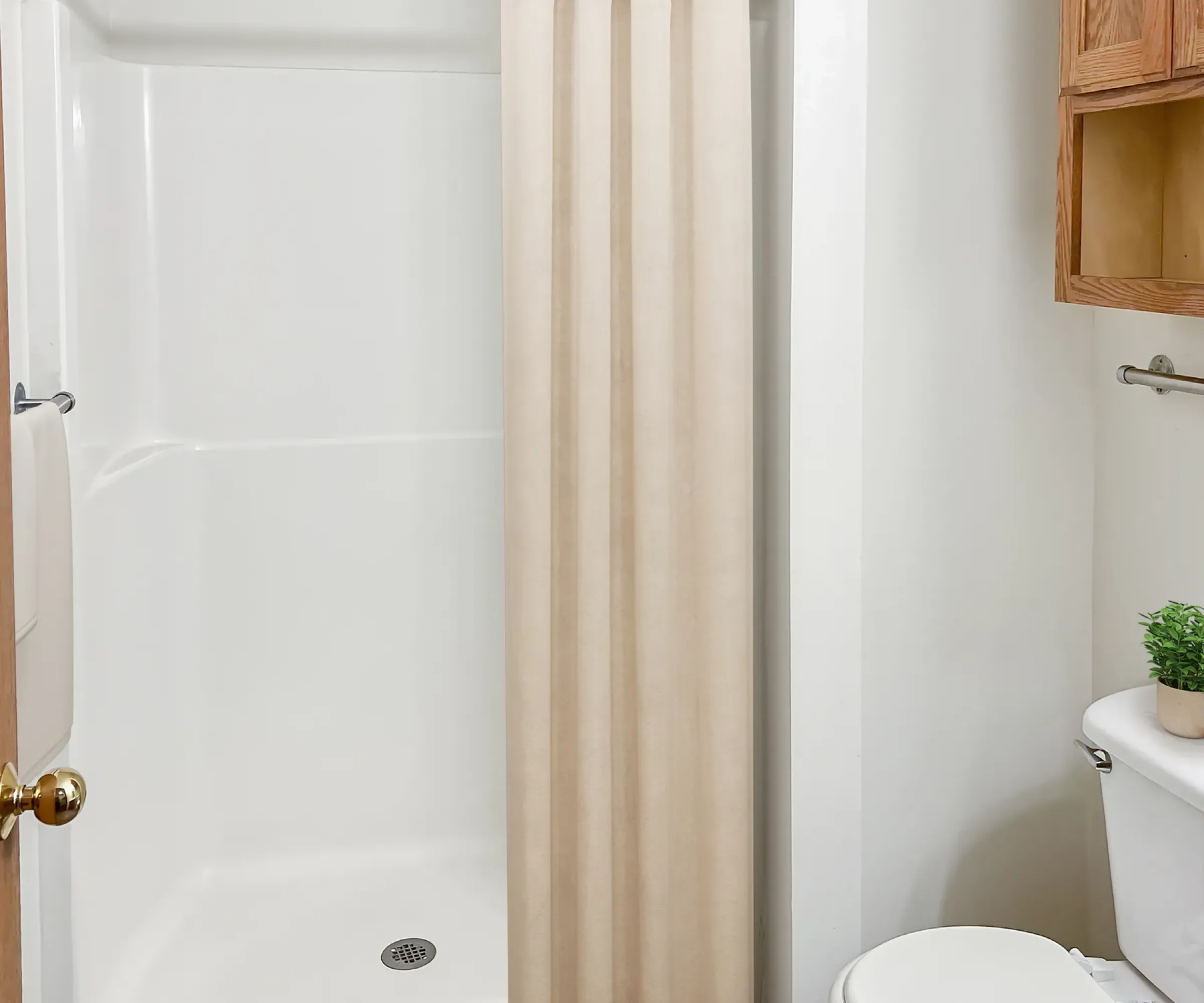 Aeon Affordable Homes and Community Affordable apartment homes with a bathroom featuring a shower, beige curtain, white toilet, wooden cabinet, and plant on a shelf.