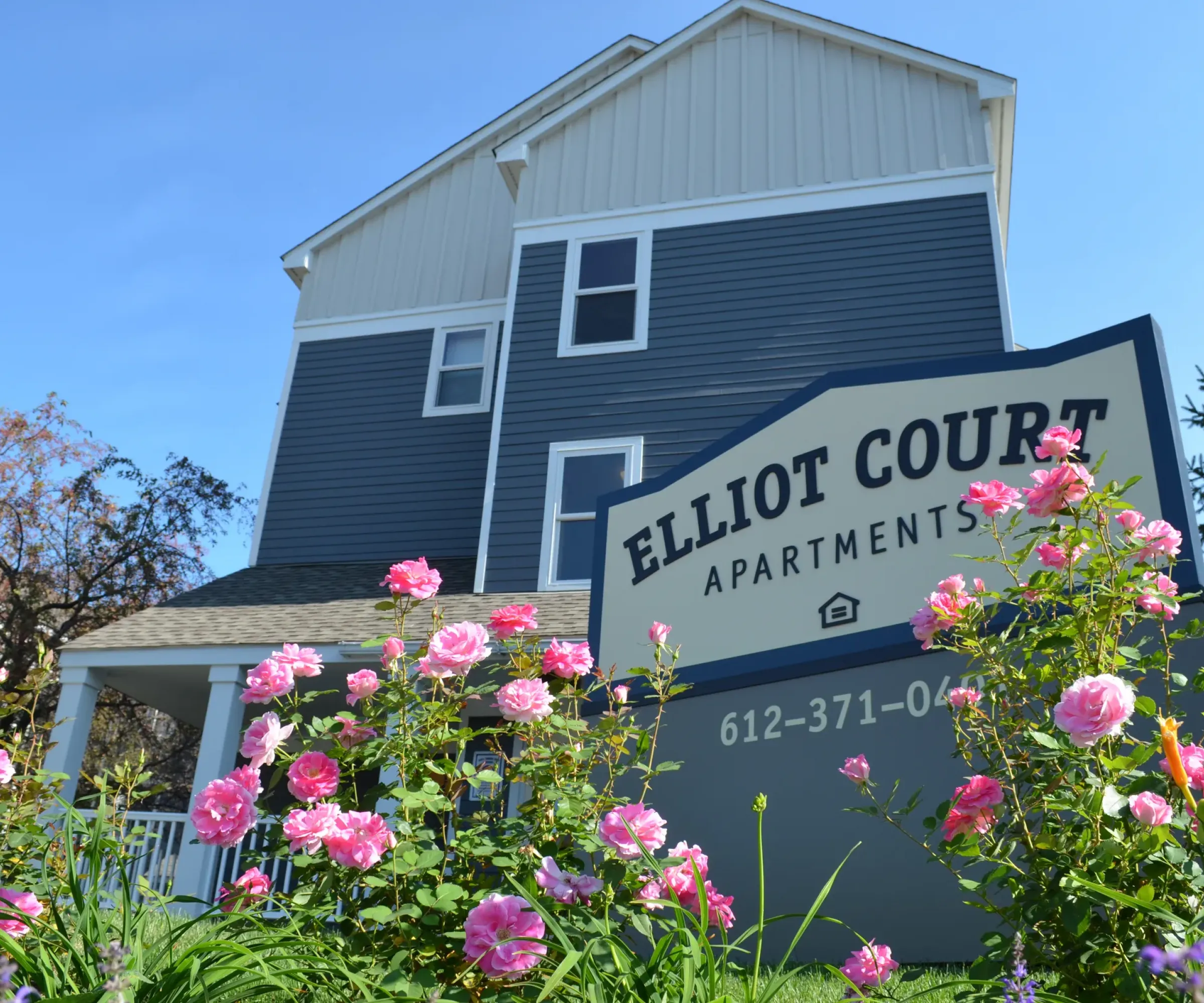 Aeon Affordable Homes and Community Sign for affordable Elliot Court Apartments in front of a blue building, surrounded by pink flowers under a clear sky.