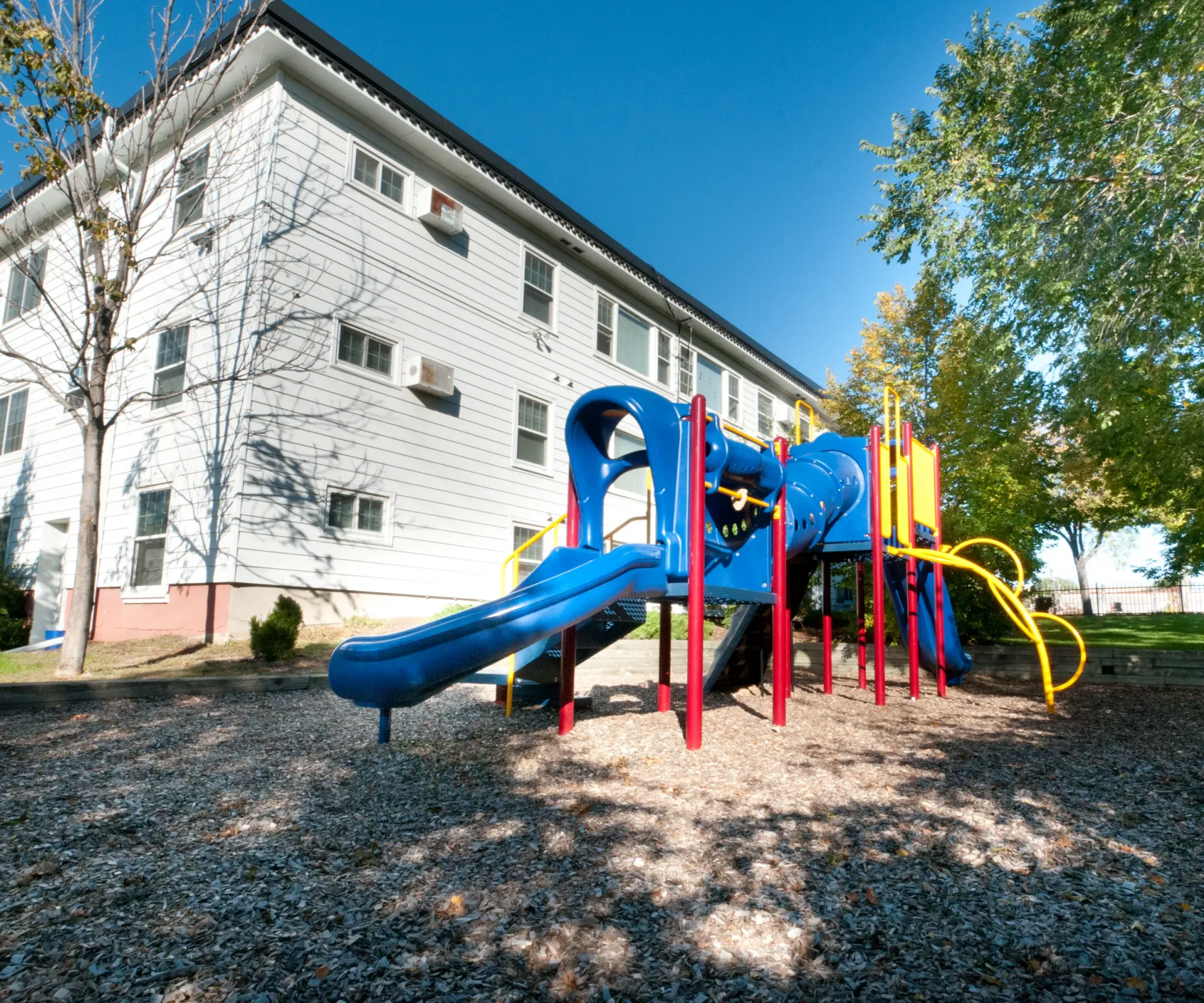 Aeon Affordable Homes and Community Playground with a blue and red slide near affordable apartment homes, surrounded by trees under a clear blue sky.