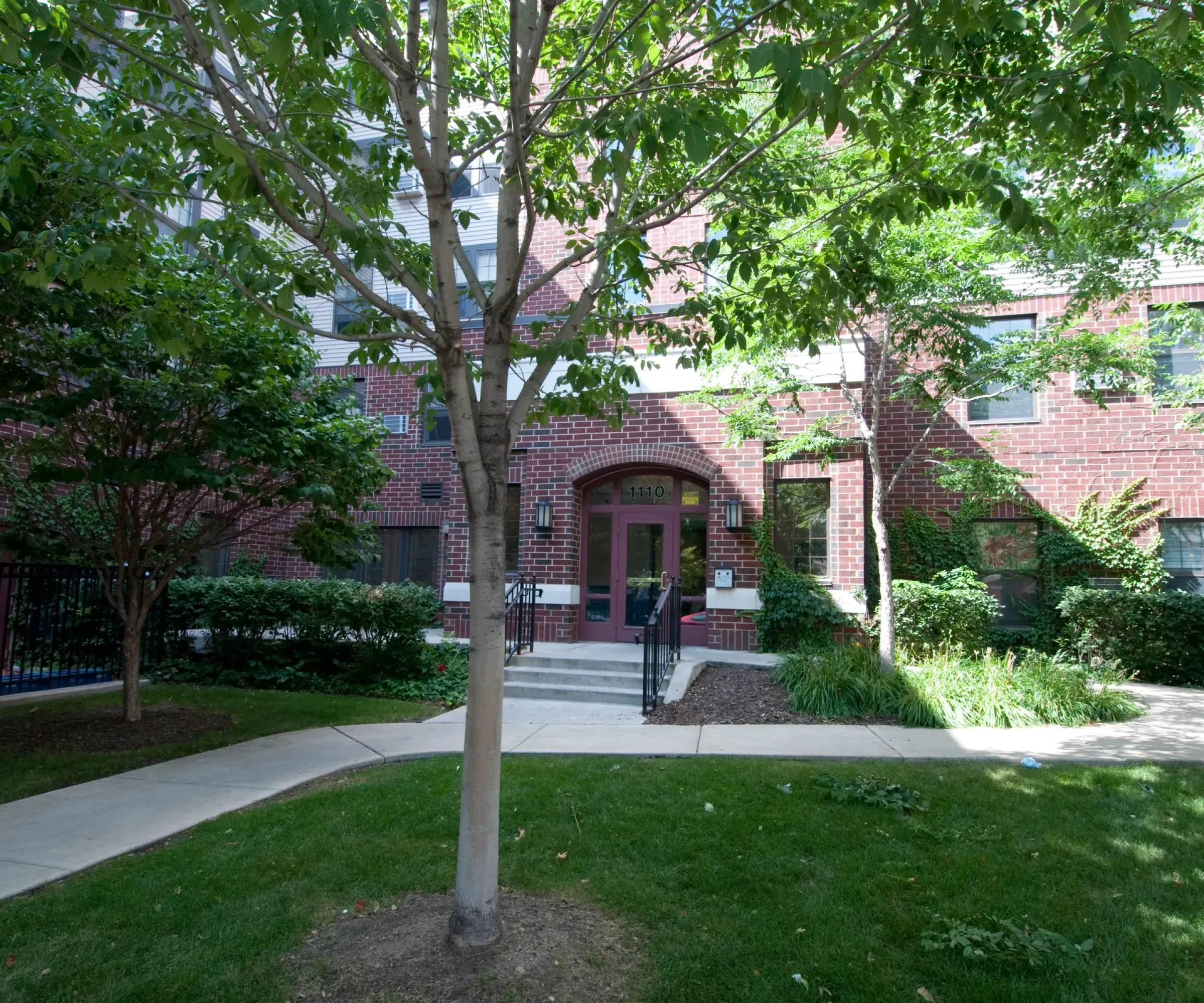 Aeon Affordable Homes and Community Entrance of red brick affordable apartment homes with green trees, grass, and a curved path leading to the door.