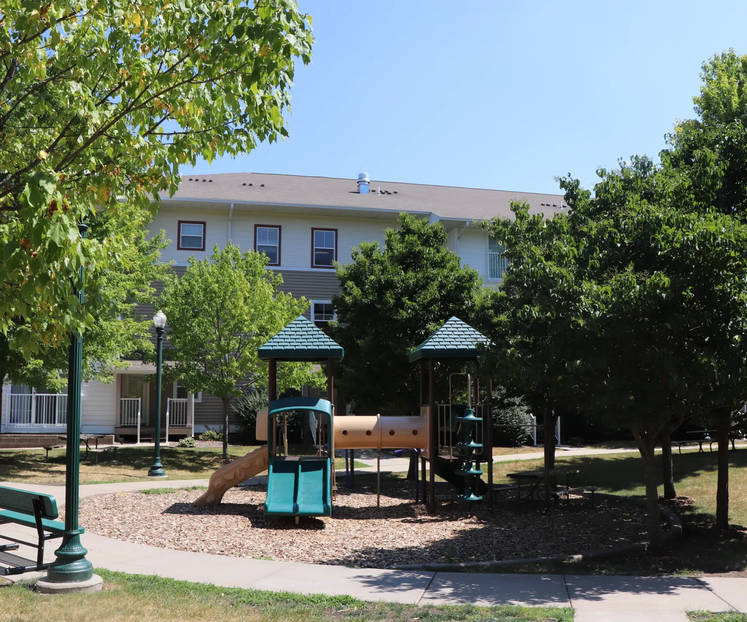 Aeon Affordable Homes and Community Playground with slide and climbing frame near affordable apartment homes, surrounded by trees and benches.