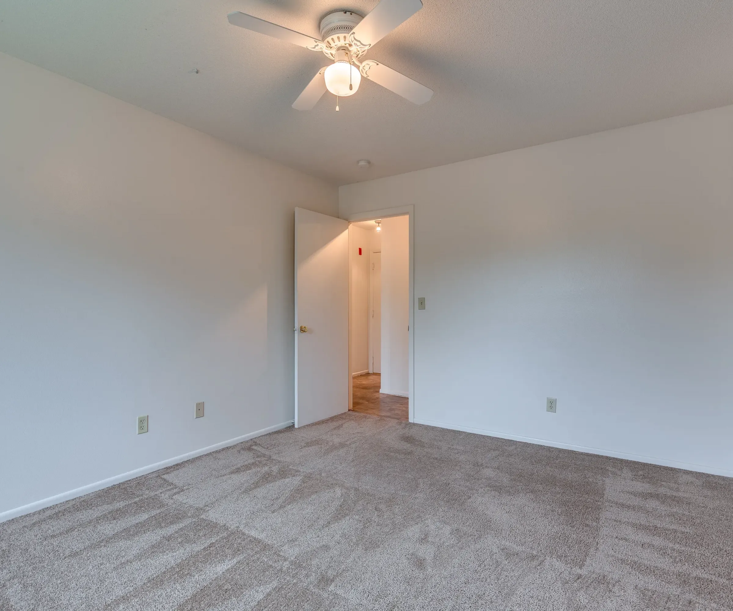 Aeon Affordable Homes and Community Affordable apartment homes featuring an empty room with beige carpet, white walls, a ceiling fan, and an open door leading to the hallway.