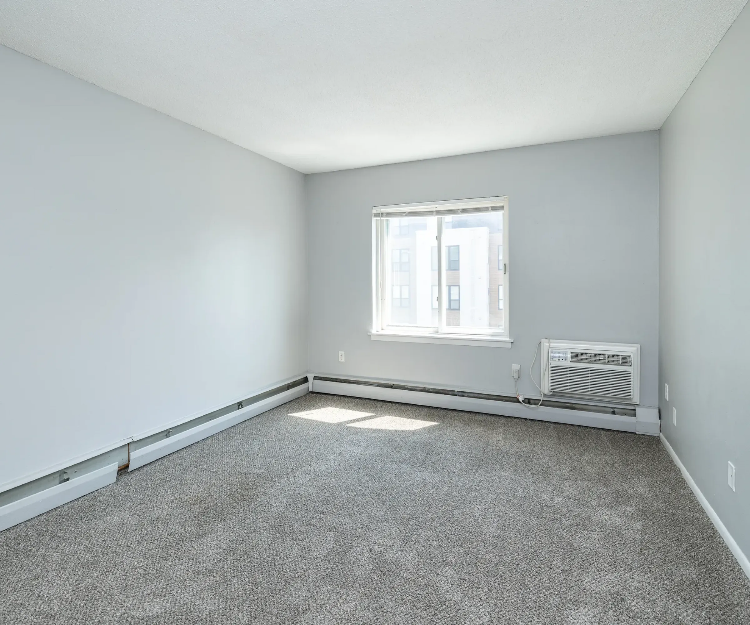 Aeon Affordable Homes The empty room at Blooming Meadows South features gray walls, a window with blinds, gray carpet, and a wall-mounted air conditioning unit.