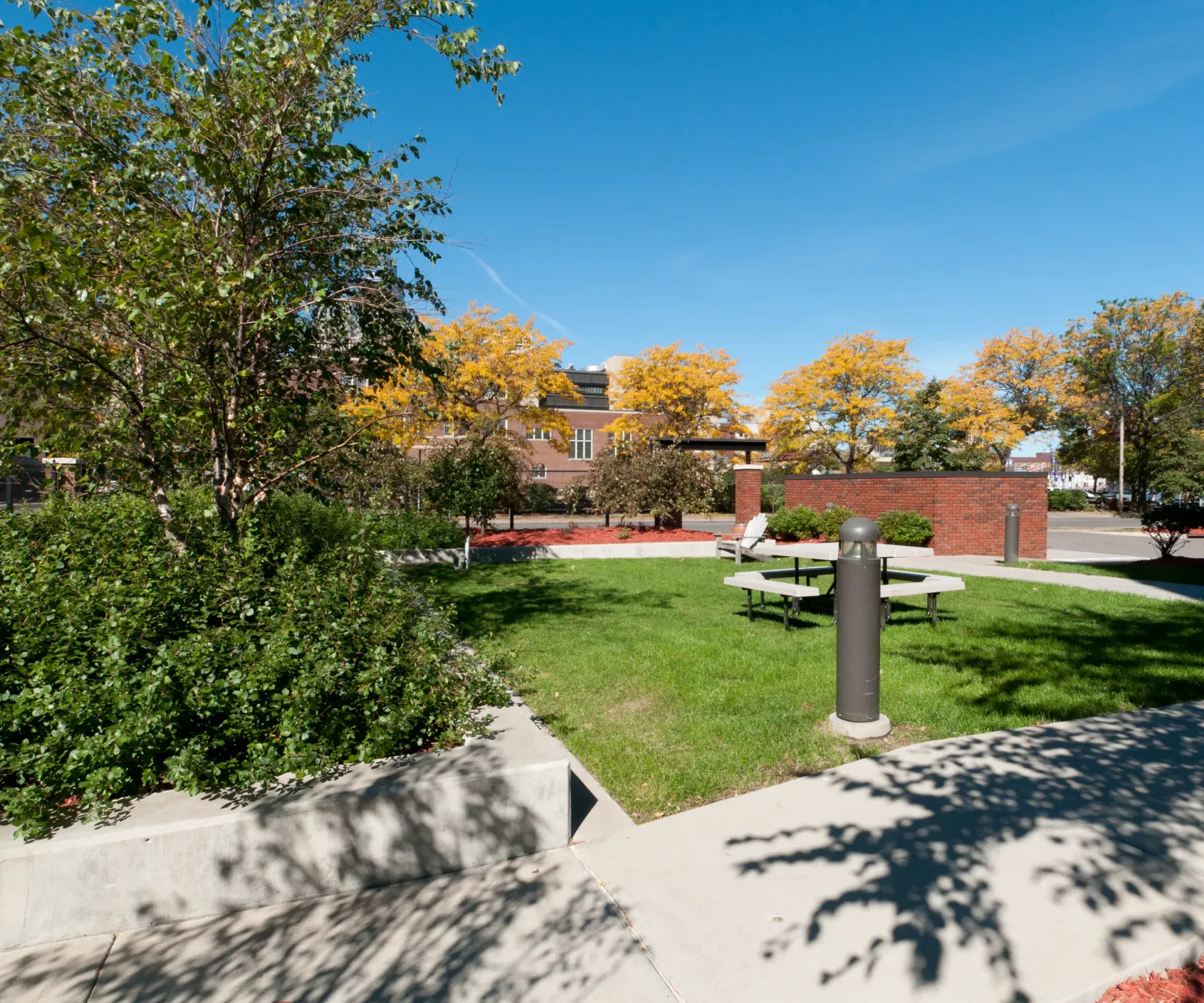 Aeon Affordable Homes and Community Sunny park scene with trees, lawn, and picnic tables. Affordable apartment homes visible in the background and a path in the foreground.