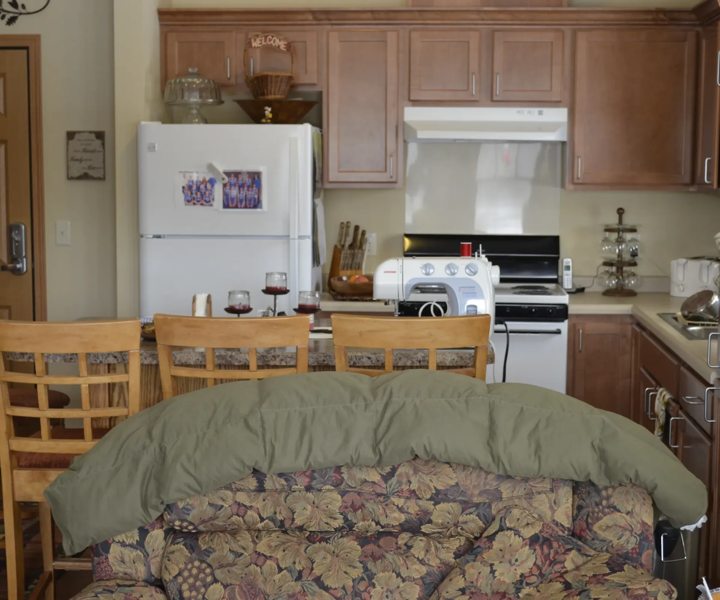 Aeon Affordable Homes and Community Cozy kitchen in affordable apartment homes with wooden cabinets, a floral couch, bar stools, and a counter with wine glasses.