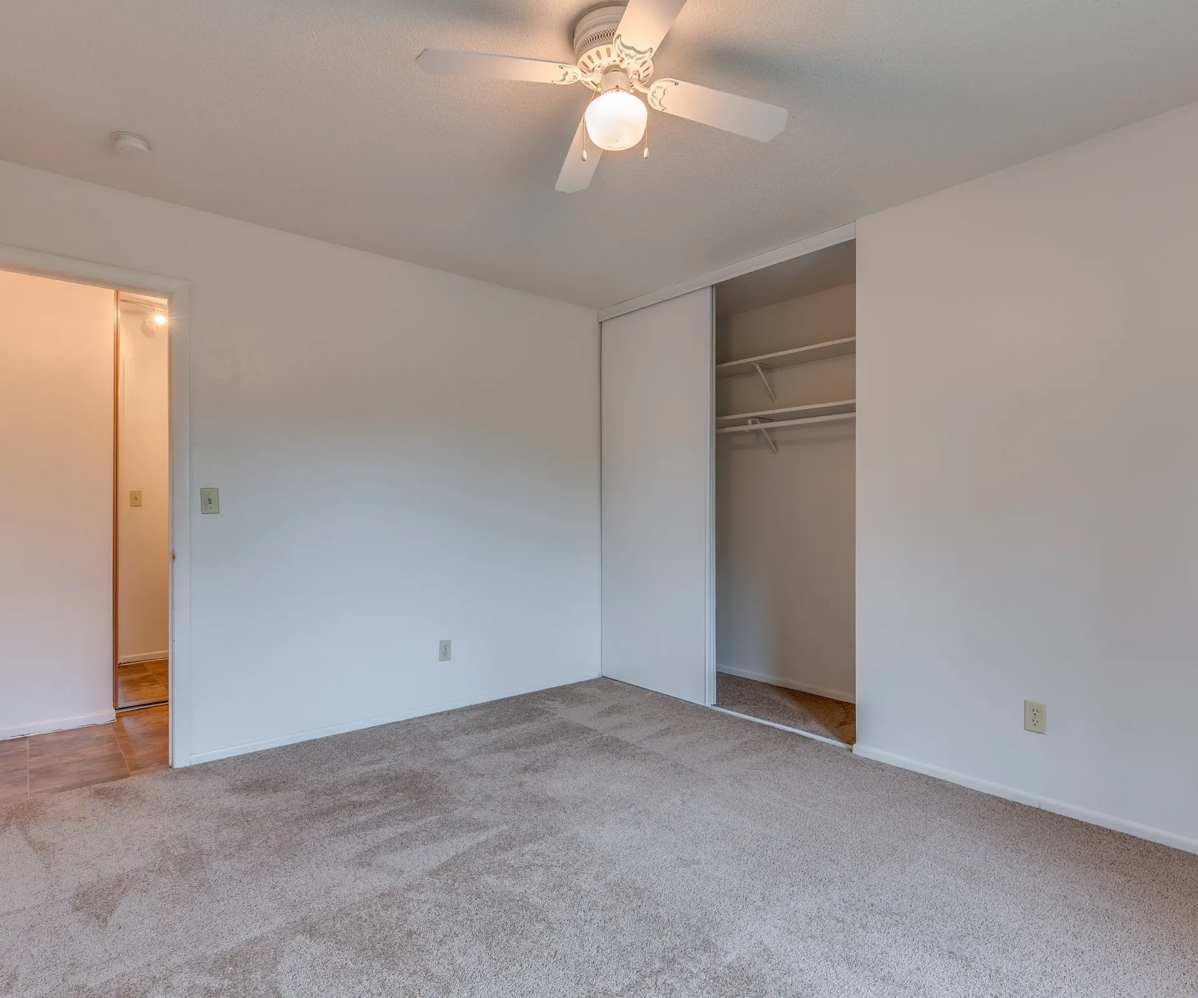 Aeon Affordable Homes and Community Affordable apartment home with an empty carpeted room, white walls, ceiling fan, open closet, and an open door leading to a hallway.