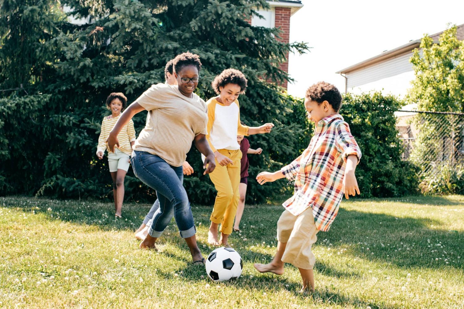 Aeon Affordable Homes Group of kids playing soccer and having fun on a grassy field with trees and houses in the background.