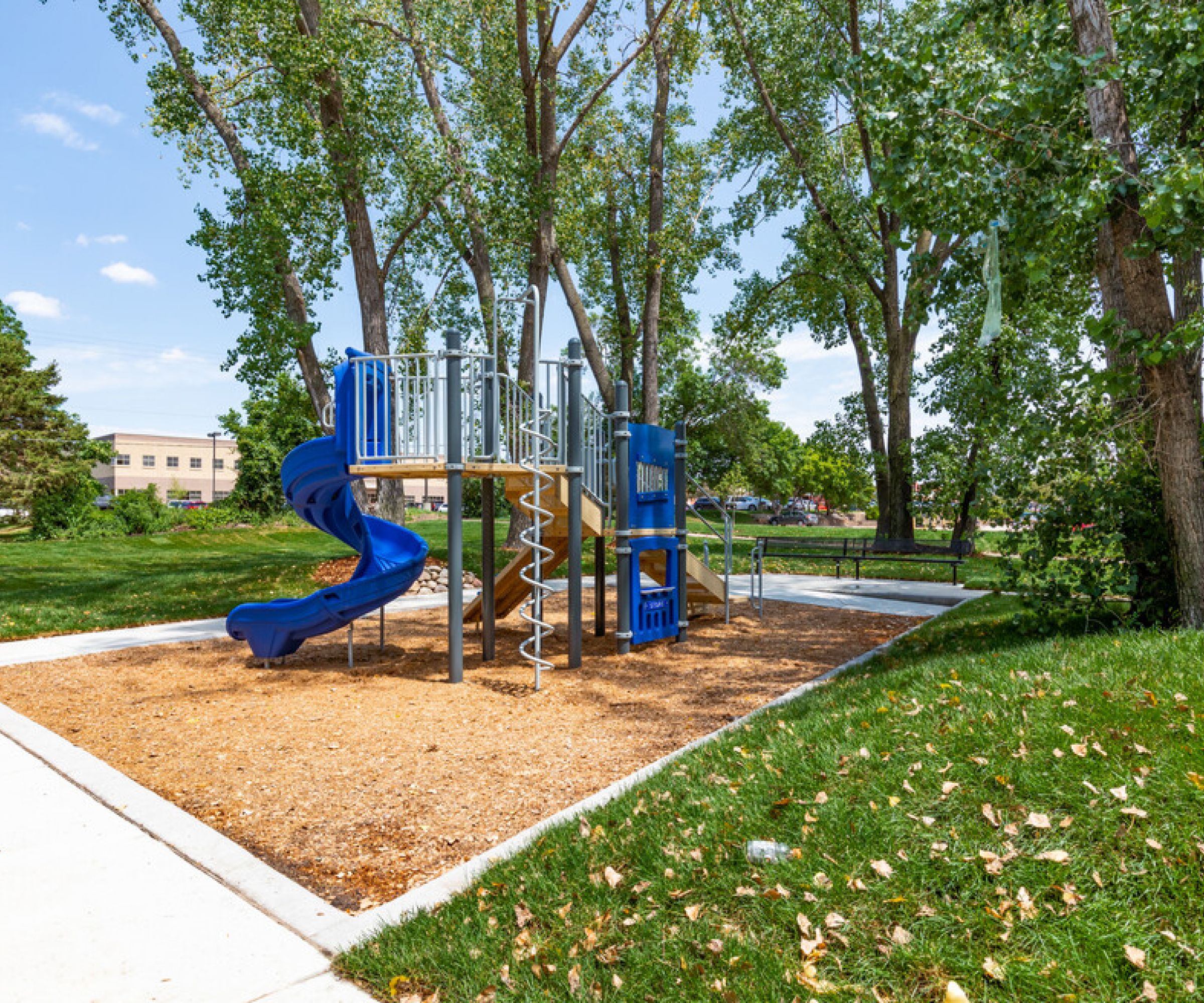 Aeon Affordable Homes and Community Playground with blue slides, metal climbing structure, and wood chips surface, nestled among trees near affordable apartment homes.
