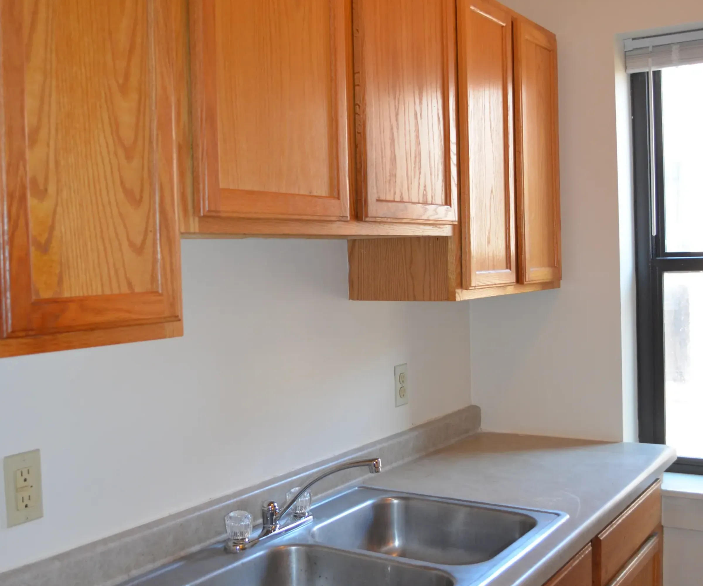 Aeon Affordable Homes and Community A cozy kitchen in affordable apartment homes, featuring wooden cabinets, a double stainless steel sink, and a countertop below a window.