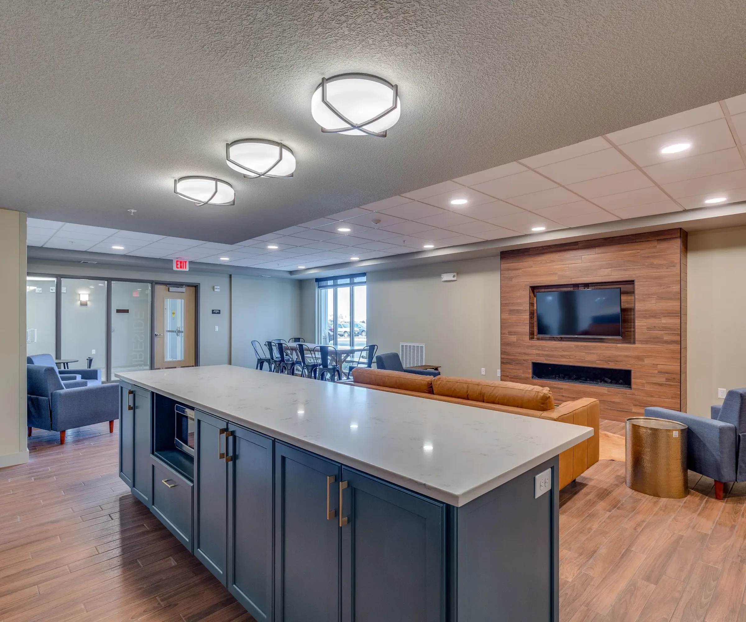 Aeon Affordable Homes and Community Modern lounge in affordable apartment homes with a kitchen island, seating area, TV, and wood flooring under soft ceiling lights.