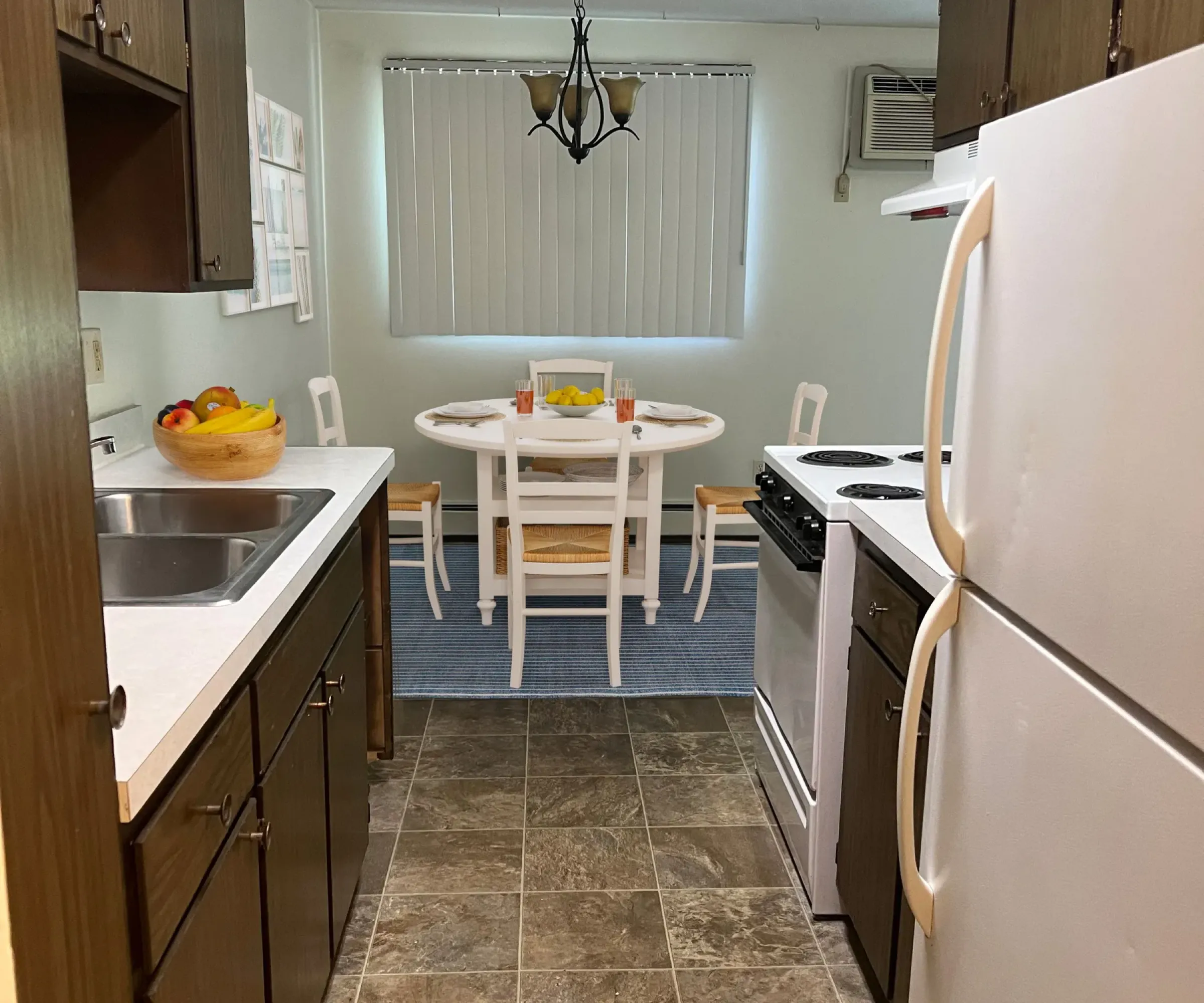 Aeon Affordable Homes and Community Compact kitchen with dark wood cabinets, stove, and fridge in affordable apartment homes. Dining area with a table and chairs visible in the background.