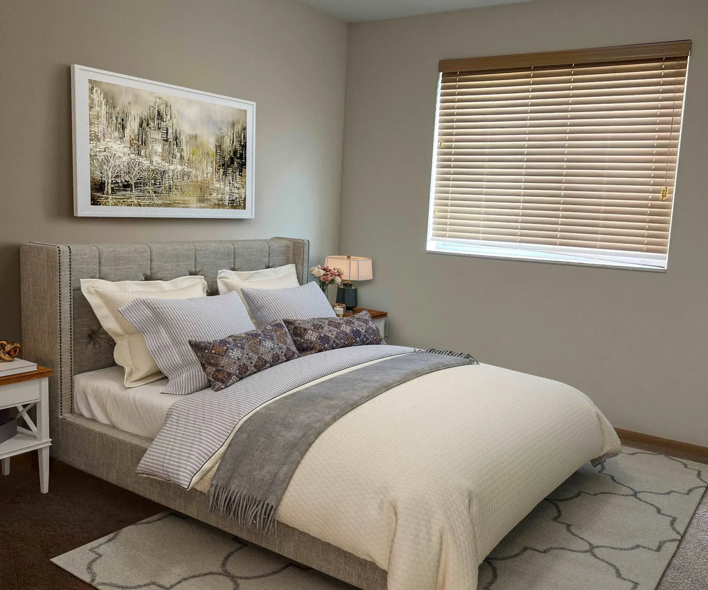 Aeon Affordable Homes and Community Cozy bedroom in affordable apartment homes, featuring a cushioned bed, decorative pillows, lamps on nightstands, artwork, and a window with blinds.