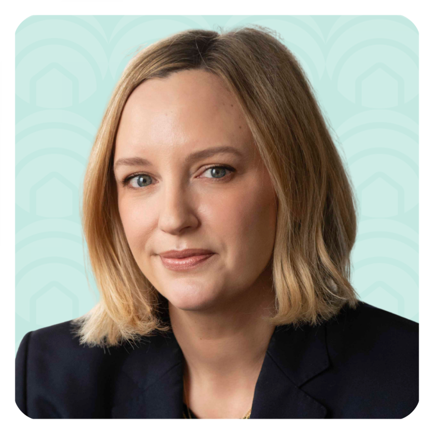 Aeon Affordable Homes A woman with blonde hair in a black blazer smiles softly at the camera against a light blue patterned background.