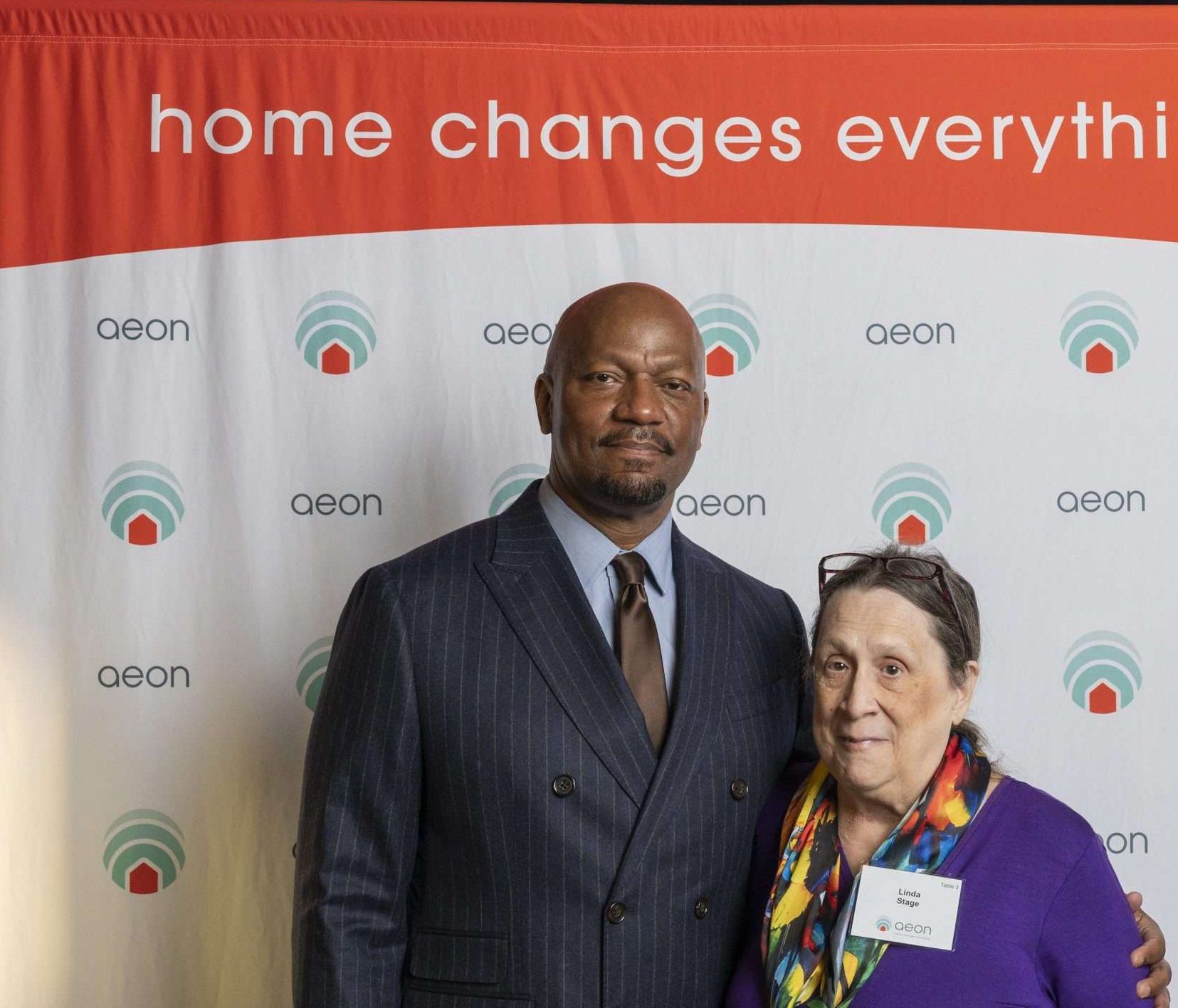 Aeon Affordable Homes and Community Two people pose in front of an "aeon" backdrop that reads "home changes everything. Aeon Affordable Homes and Community Two people posing in front of a banner with "home changes everything" text and Aeon logos. Eric Johnson and Linda Stage Eric Johnson and Linda Stage