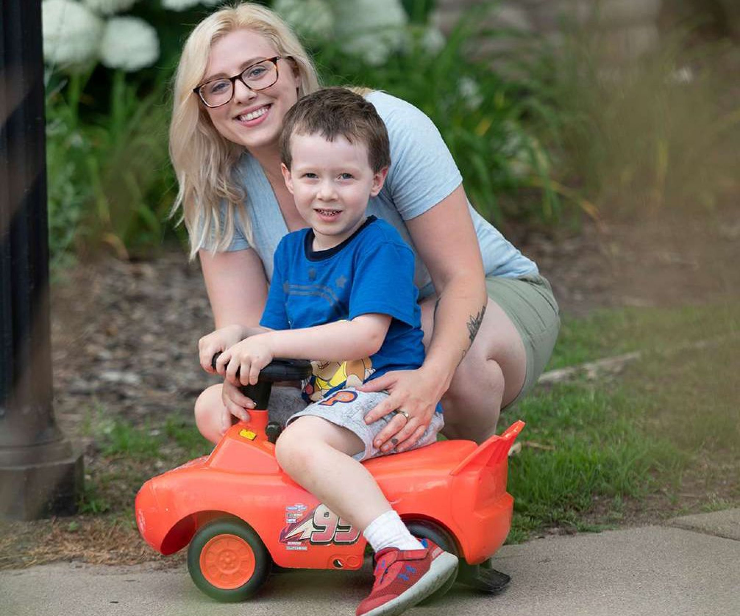 Aeon Affordable Homes Woman and child smiling; child is sitting in a small red toy car outside.