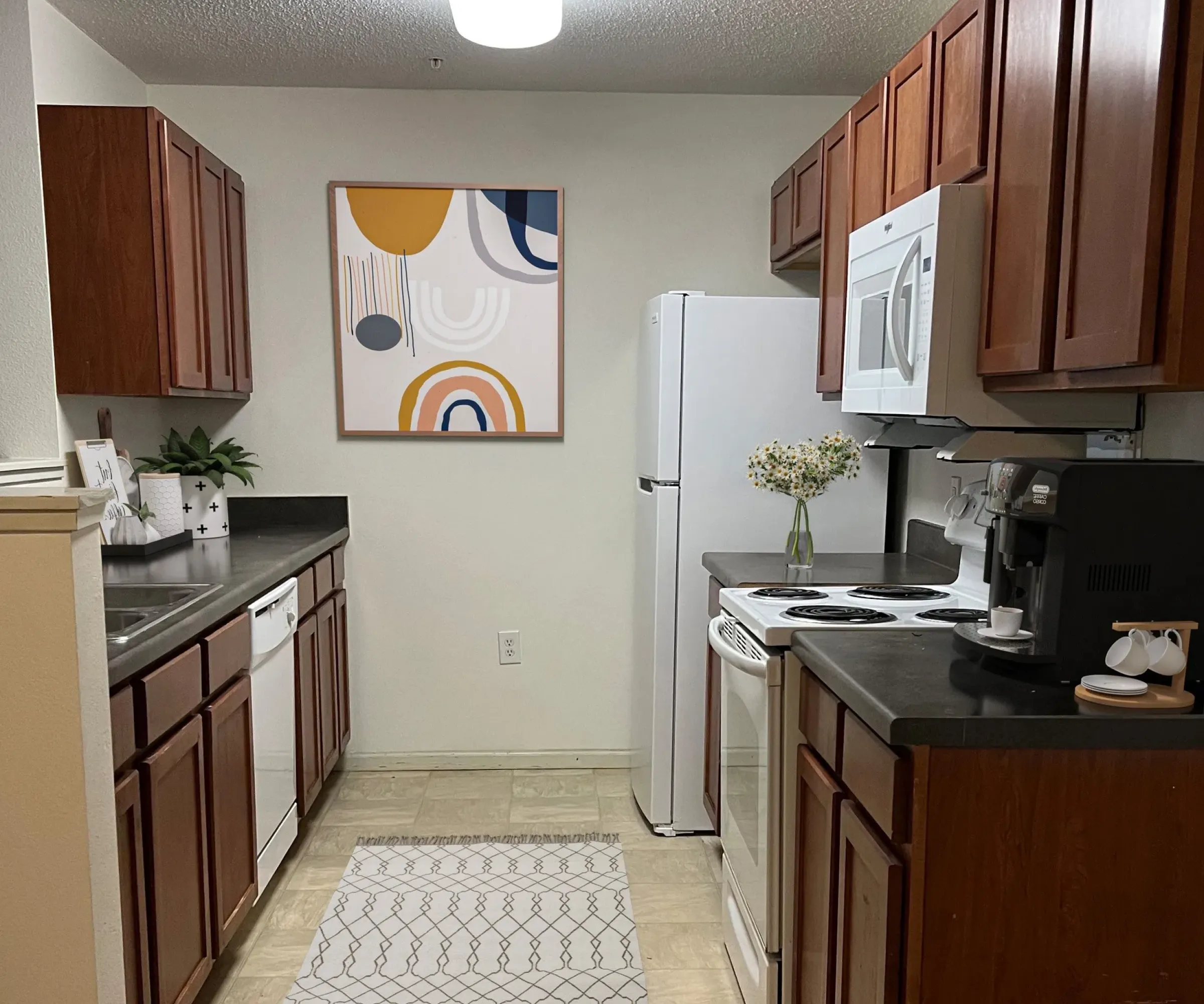 Aeon Affordable Homes and Community A cozy, affordable apartment home featuring a compact kitchen with dark countertops, white appliances, abstract art, and a patterned rug.