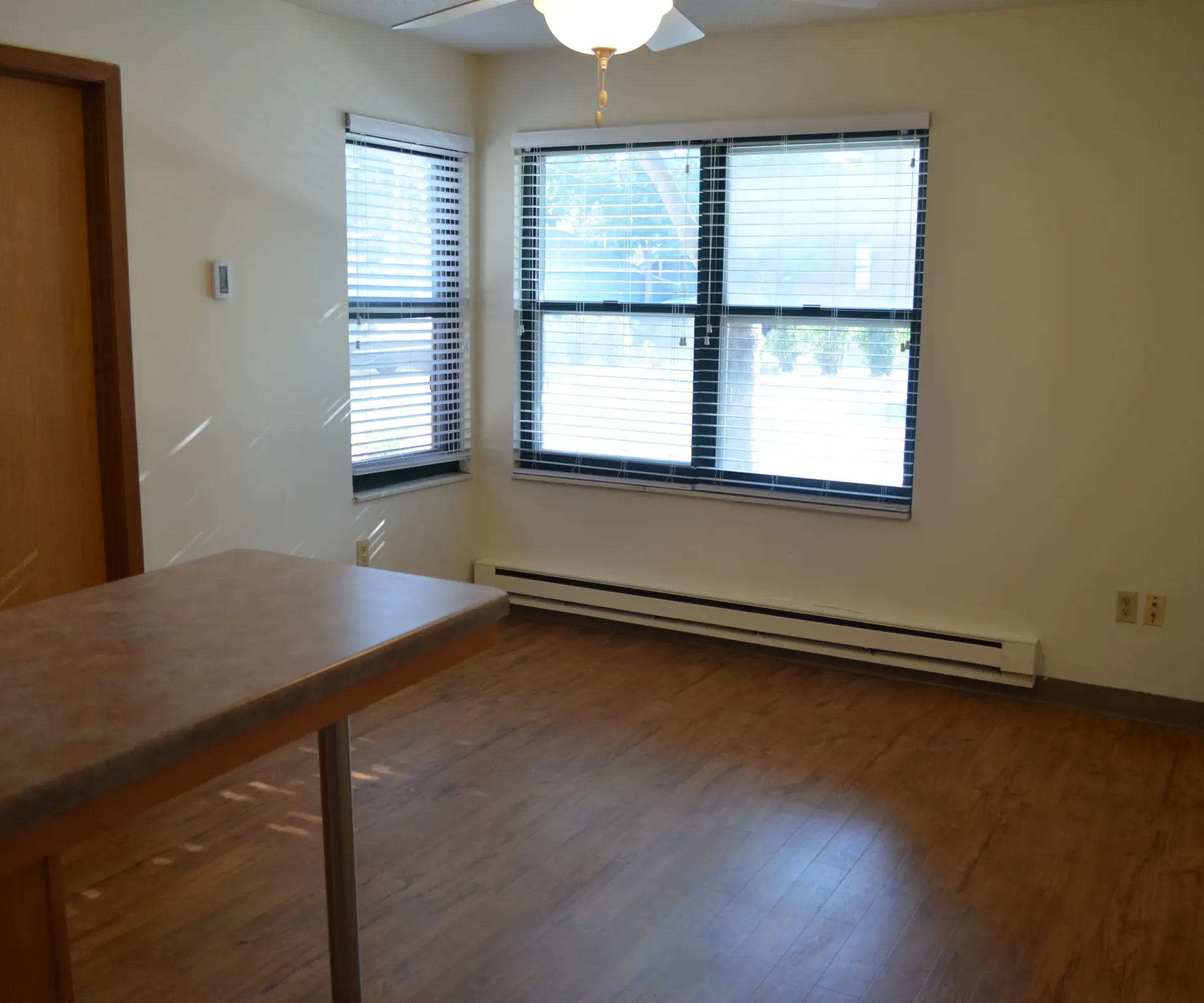 Aeon Affordable Homes and Community Empty room with wood flooring, ceiling fan, two windows with blinds, wooden door, and countertop; perfect for those seeking affordable apartment homes.
