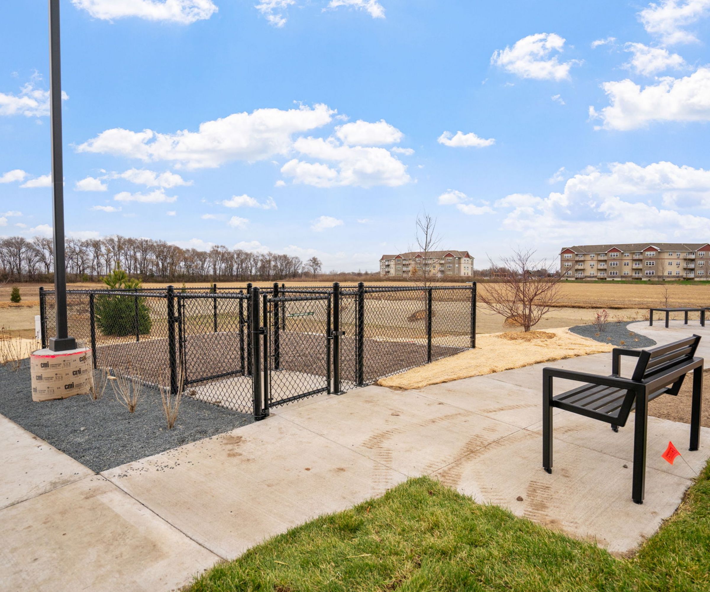 Aeon Affordable Homes and Community Fenced dog park area with seating, surrounded by pathways and grassy fields under a partly cloudy sky near affordable apartment homes.
