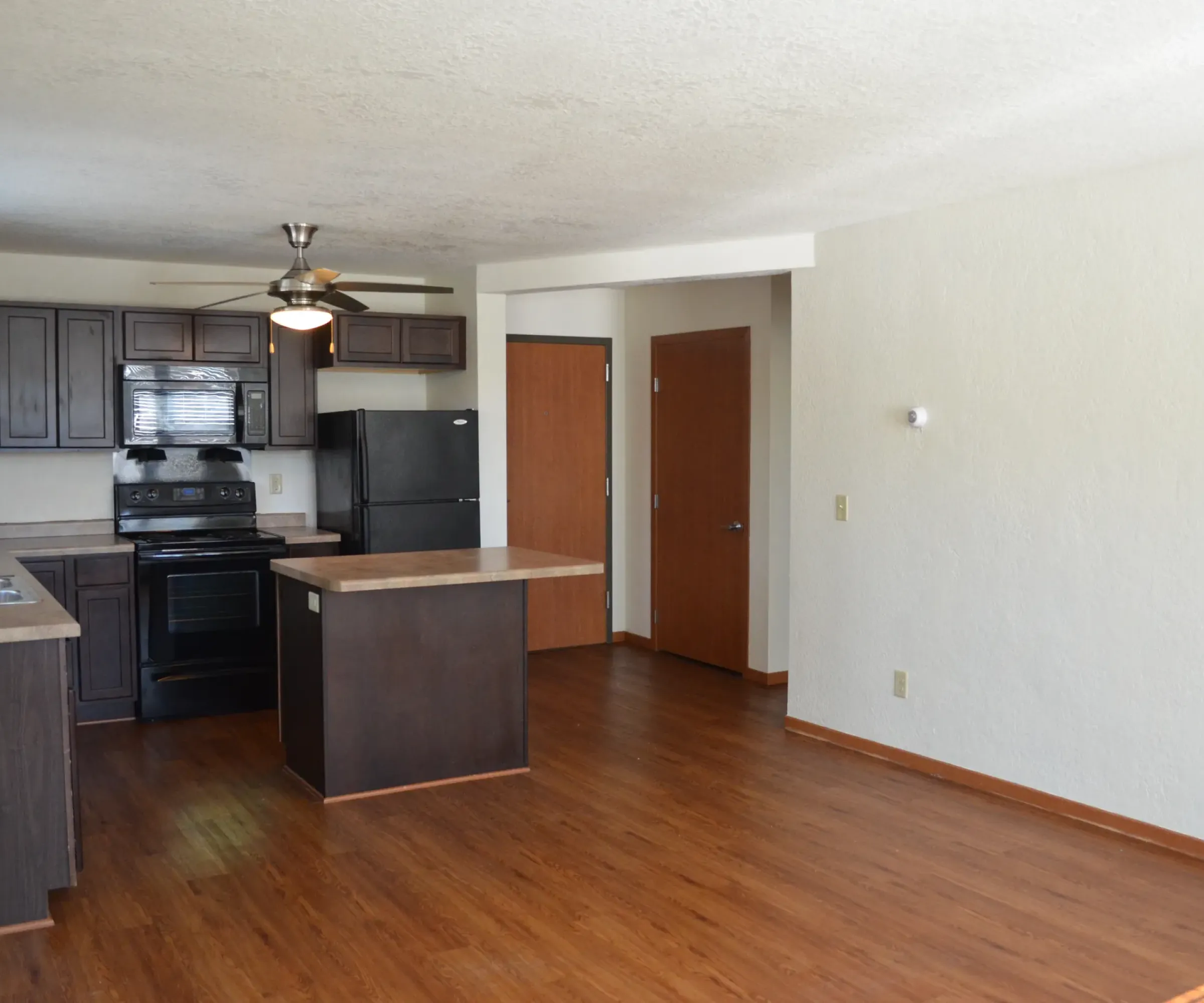 Aeon Affordable Homes and Community Open-plan kitchen in affordable apartment homes, featuring dark wood cabinets, black appliances, and an island, flowing into the wood-floored living space.