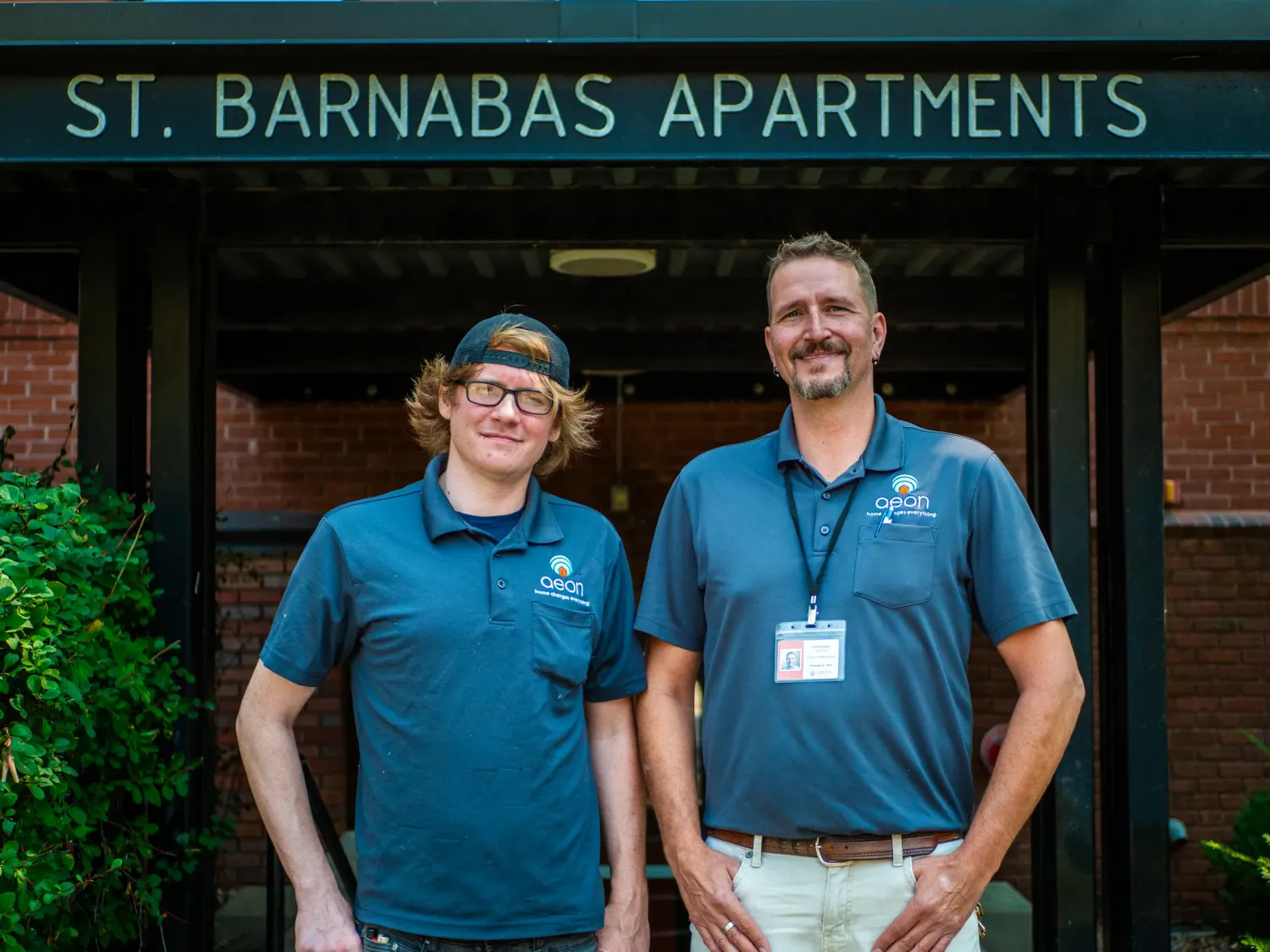 Aeon Affordable Homes Two men in matching blue shirts stand smiling in front of St. Barnabas Apartments entrance.