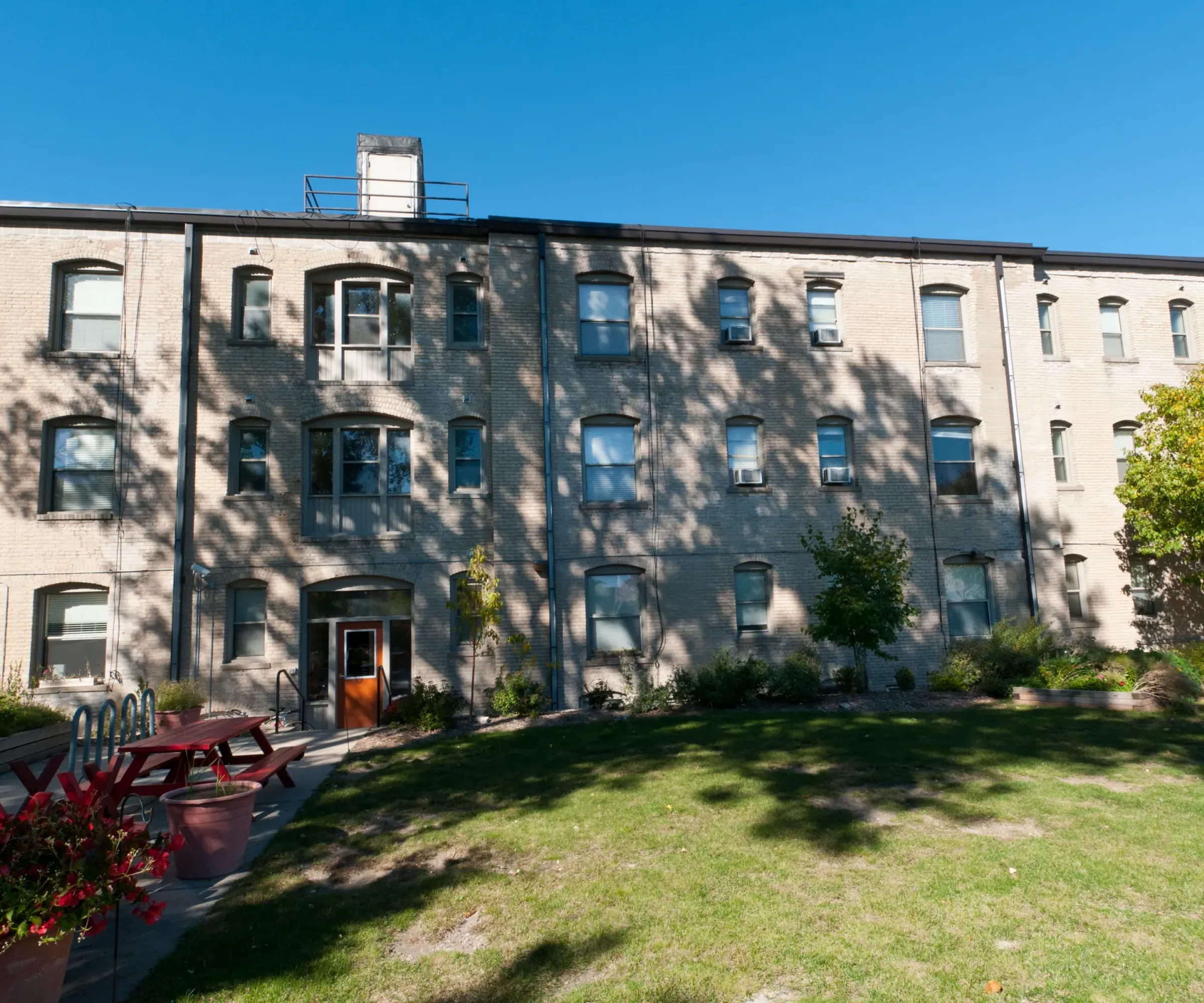 Aeon Affordable Homes and Community Affordable apartment homes in a brick building with multiple windows, a central entrance, and a courtyard with grass, benches, and tables under a clear sky.