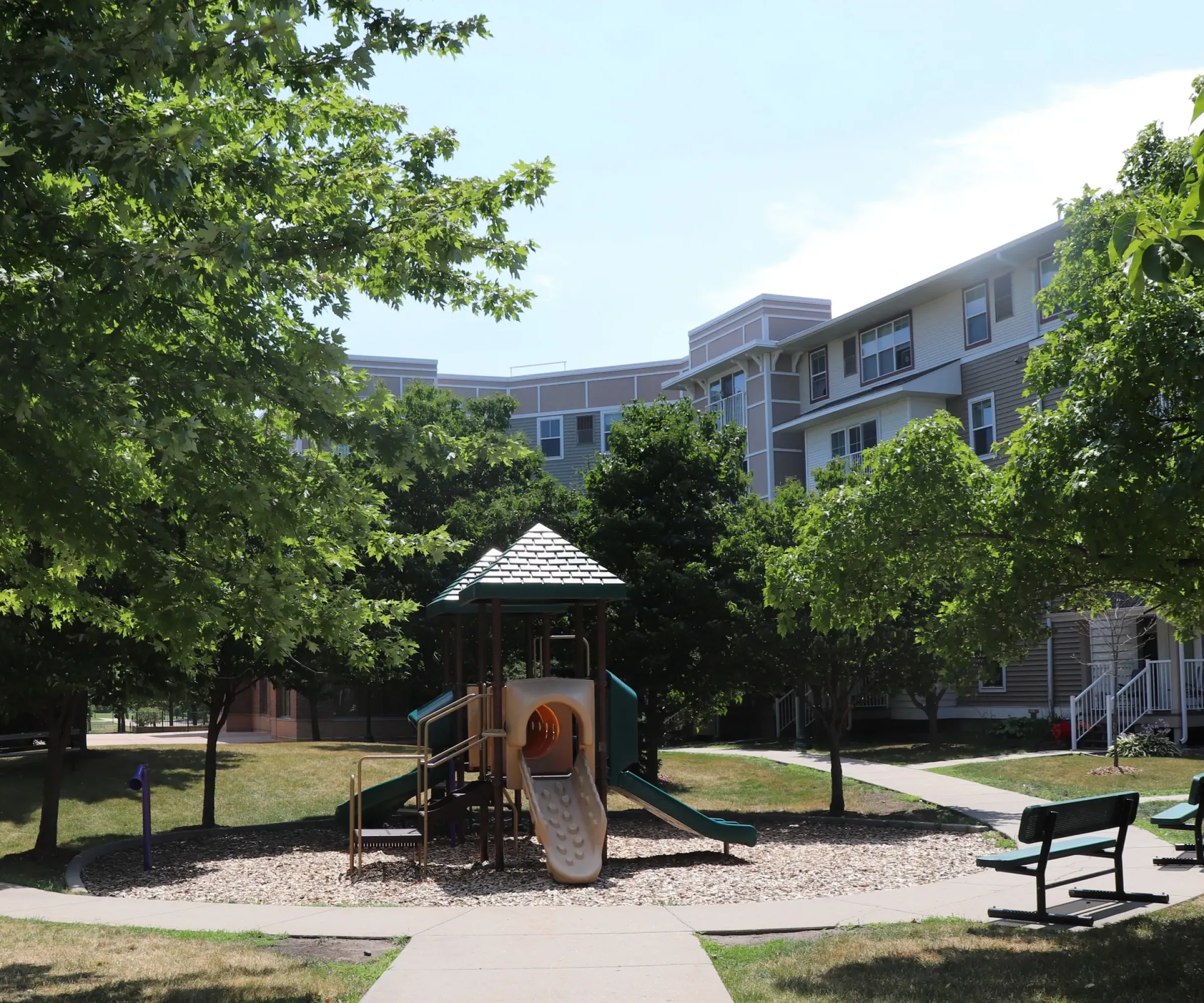 Aeon Affordable Homes and Community Playground with a slide, nestled among trees and benches, surrounded by residential buildings and affordable apartment homes under a clear sky.