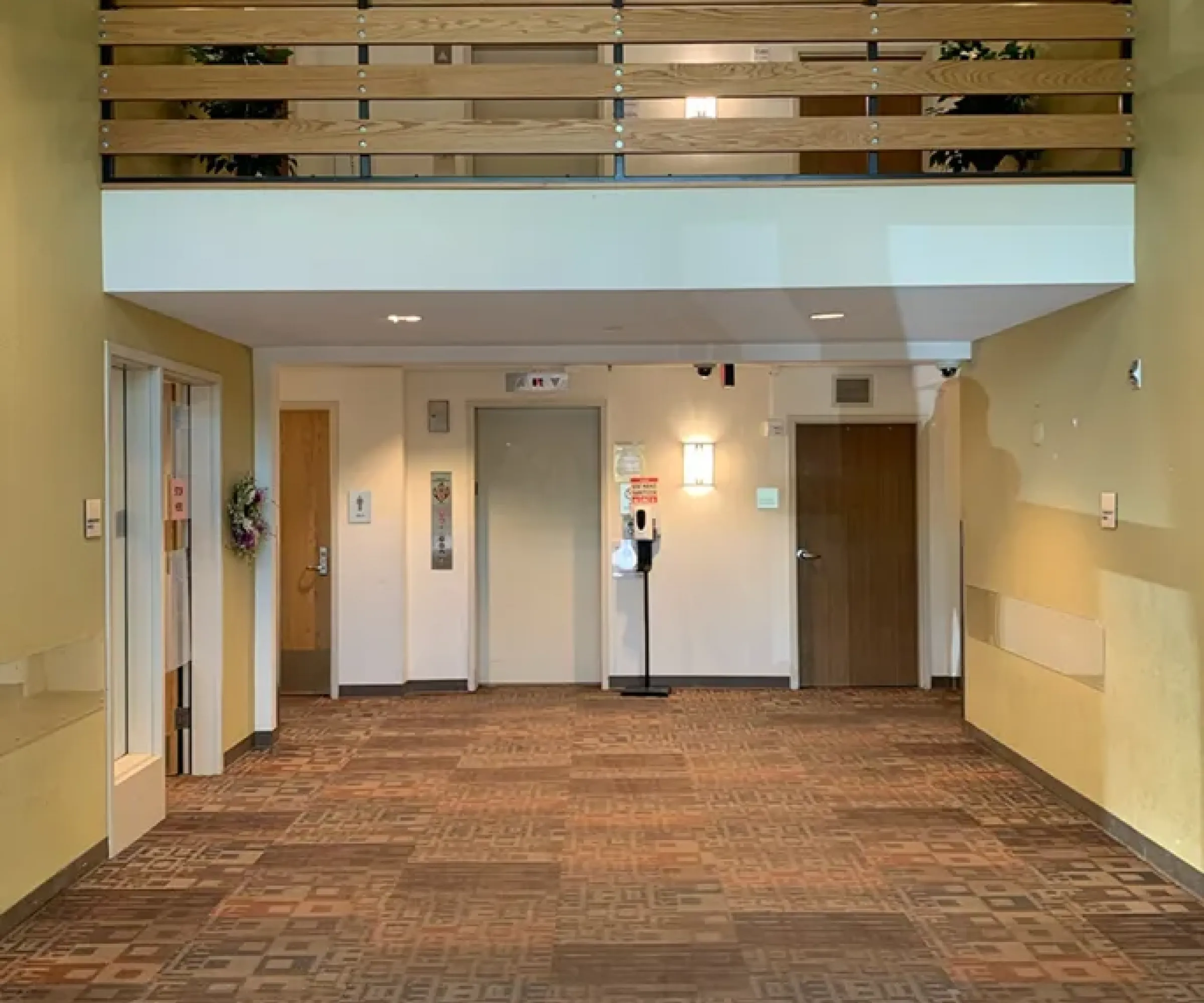 Aeon Affordable Homes and Community A hallway with light yellow walls, an elevator, a staircase, and a balcony with wooden railing above in affordable apartment homes.