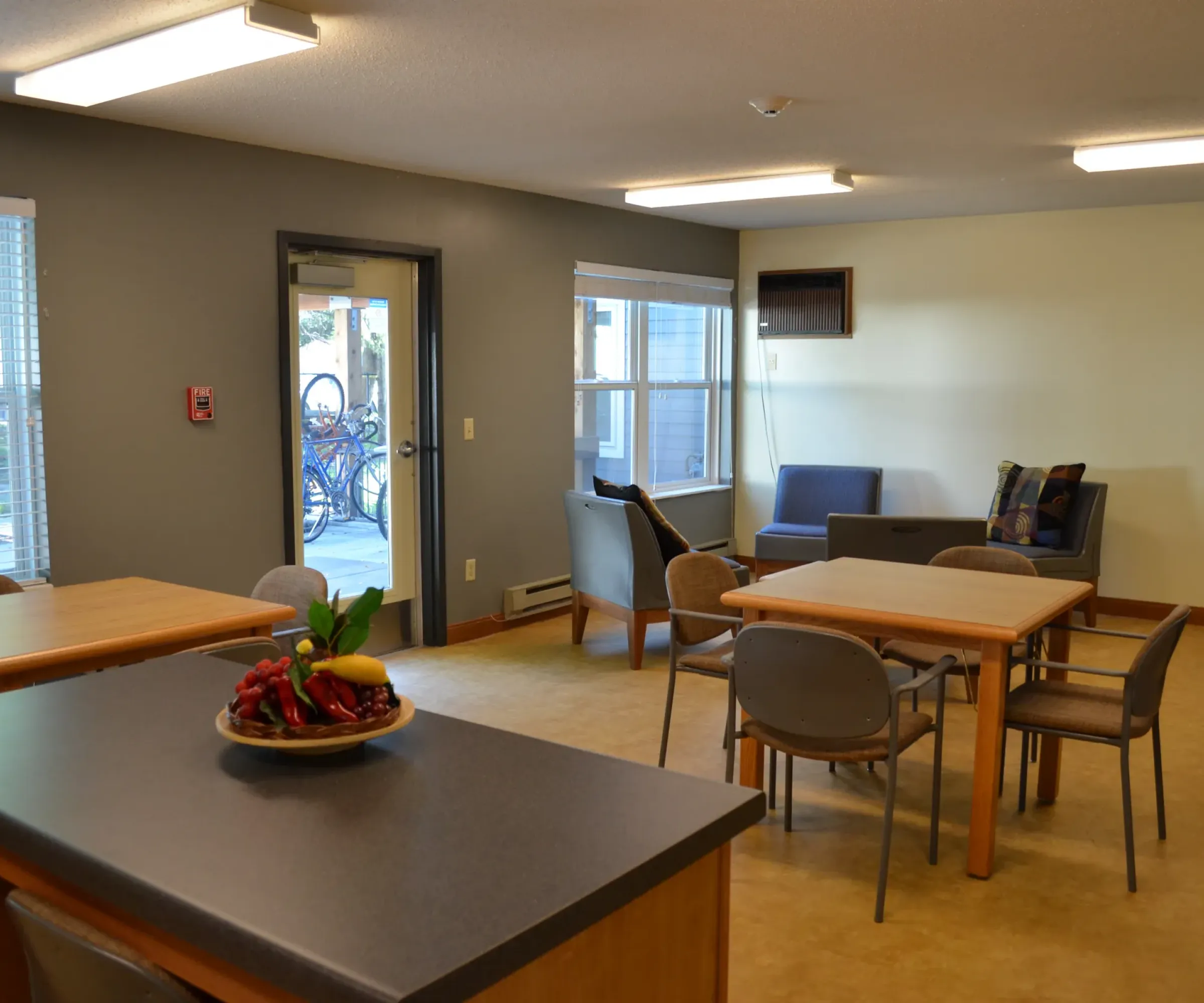 Aeon Affordable Homes and Community A cozy communal room in our affordable apartment homes features tables, chairs, a fruit countertop, and windows letting in natural light.
