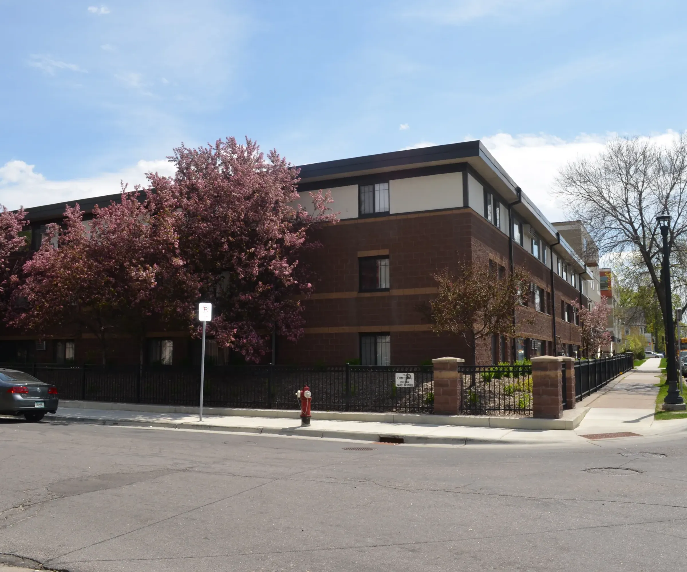 Aeon Affordable Homes and Community Affordable apartment homes in a brick building with blooming trees on a street corner, cars parked nearby under a partly cloudy sky.