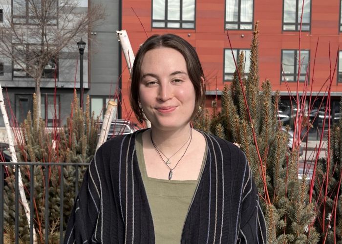 Aeon Affordable Homes A person in a striped cardigan stands outside in front of red and gray buildings on a sunny day.
