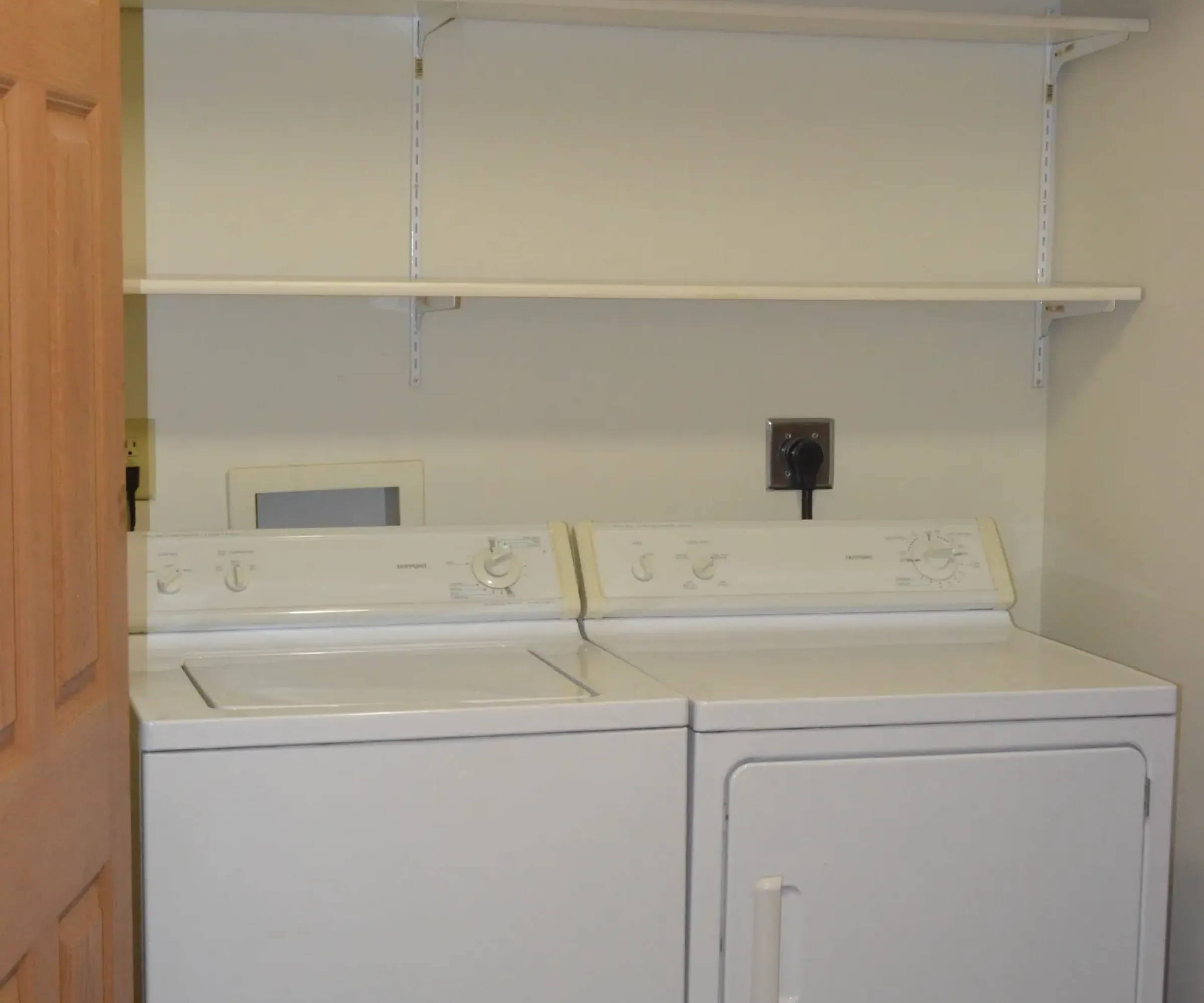 Aeon Affordable Homes and Community Laundry room in affordable apartment homes with a washer and dryer under two empty shelves, door partially open on the left.