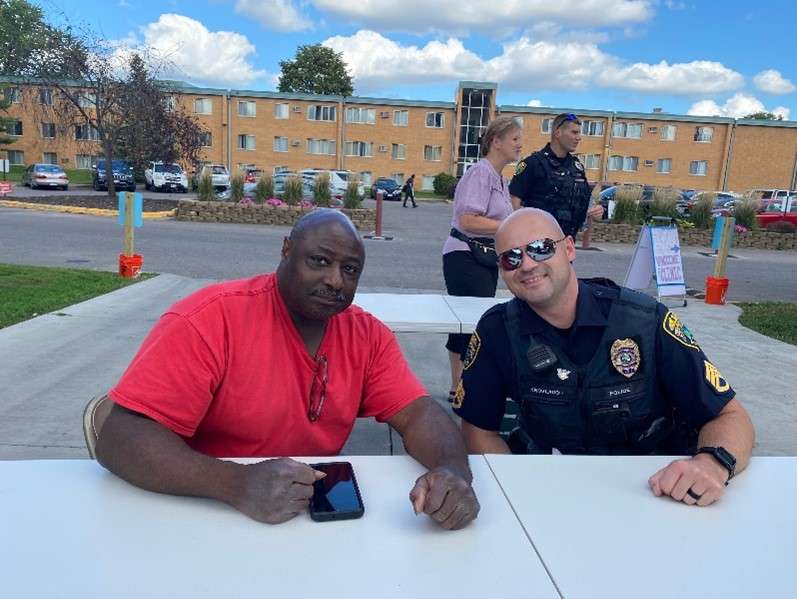 Aeon Affordable Homes and Community Two men seated at a table outdoors, one in a red shirt and the other in a police uniform, with buildings in the background.