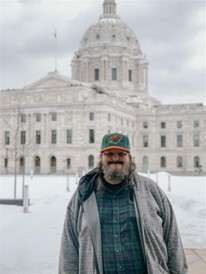 Aeon Affordable Homes and Community Man in a jacket and cap stands in front of a large, domed government building on a snowy day.