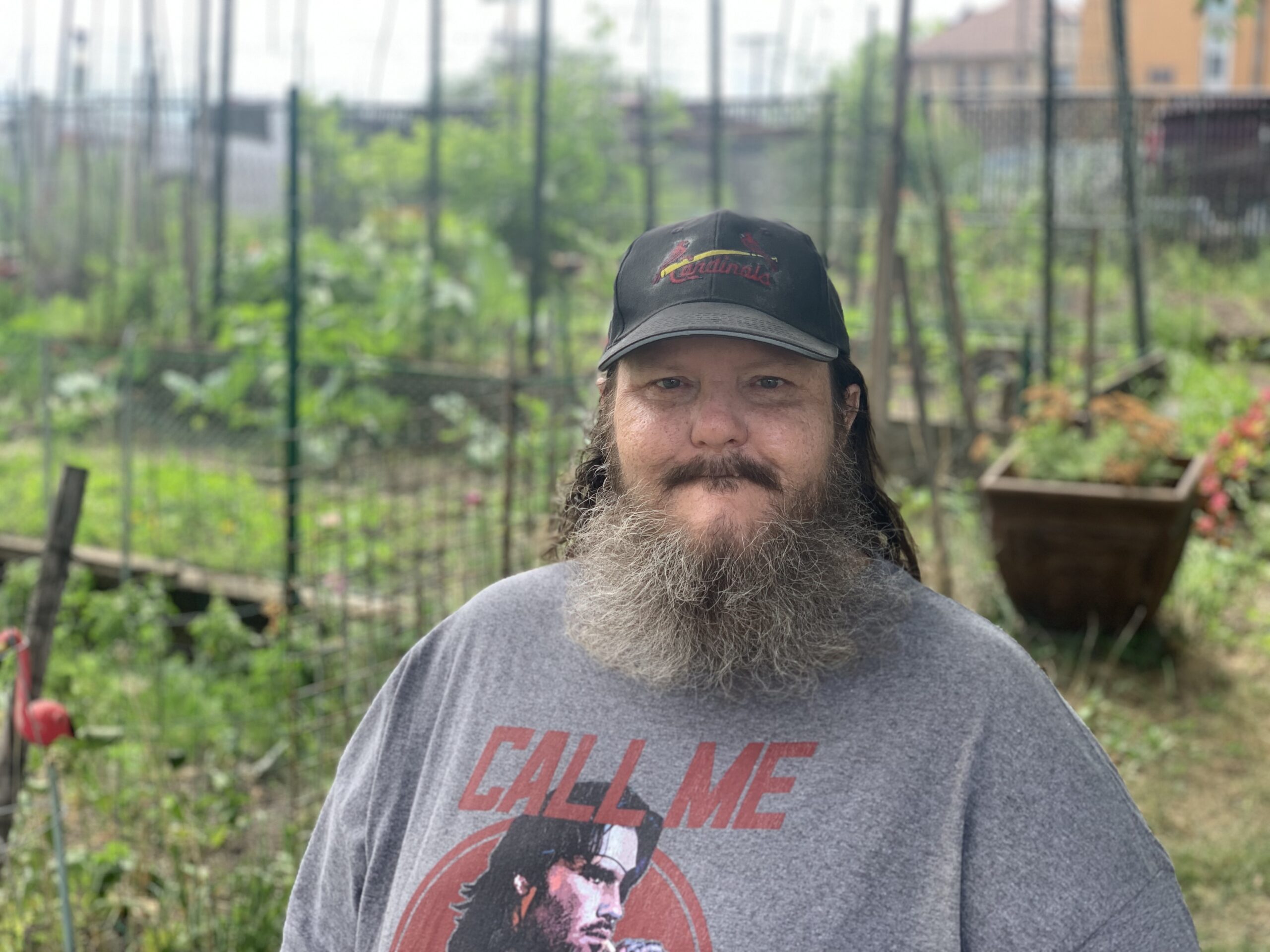 Aeon Affordable Homes Bearded man in a cap and graphic t-shirt stands in a lush garden, the perfect setting for Randy's Story among plants and trellises.